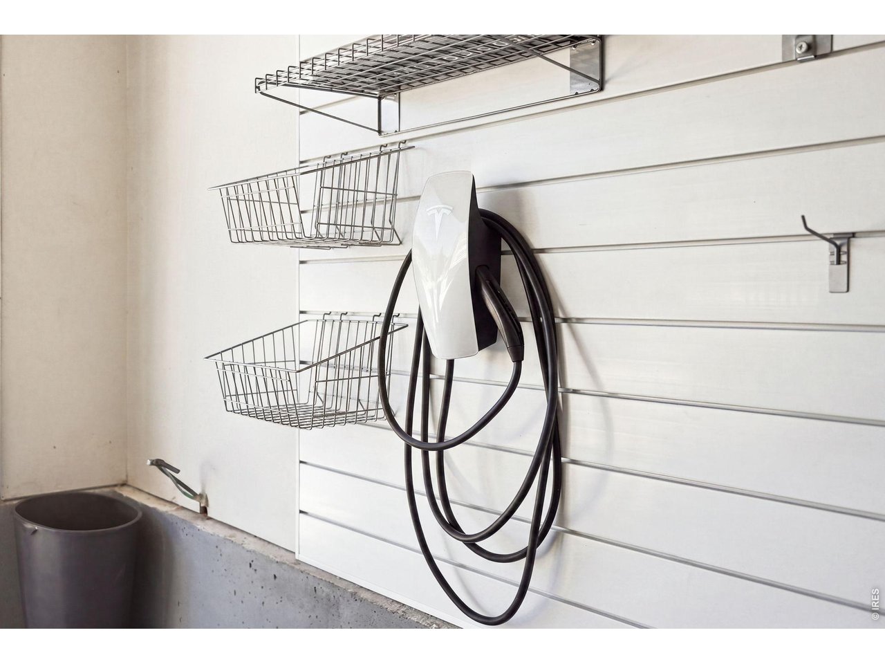 Garage Tesla charger mounted on a white slatwall beside wire storage baskets.