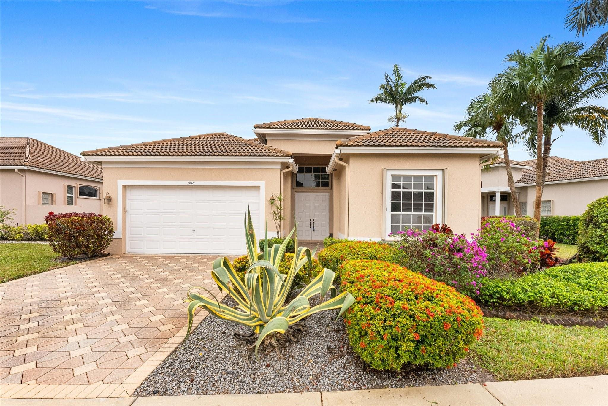 Single-story tan home with a tile roof, white garage door, paved driveway, and tropical landscaping.