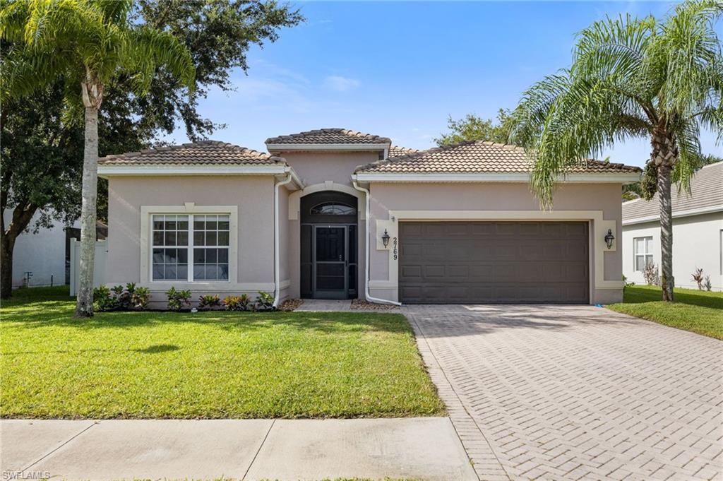 Single-story stucco home with a tiled roof, two-car garage, paver driveway, and palm trees.