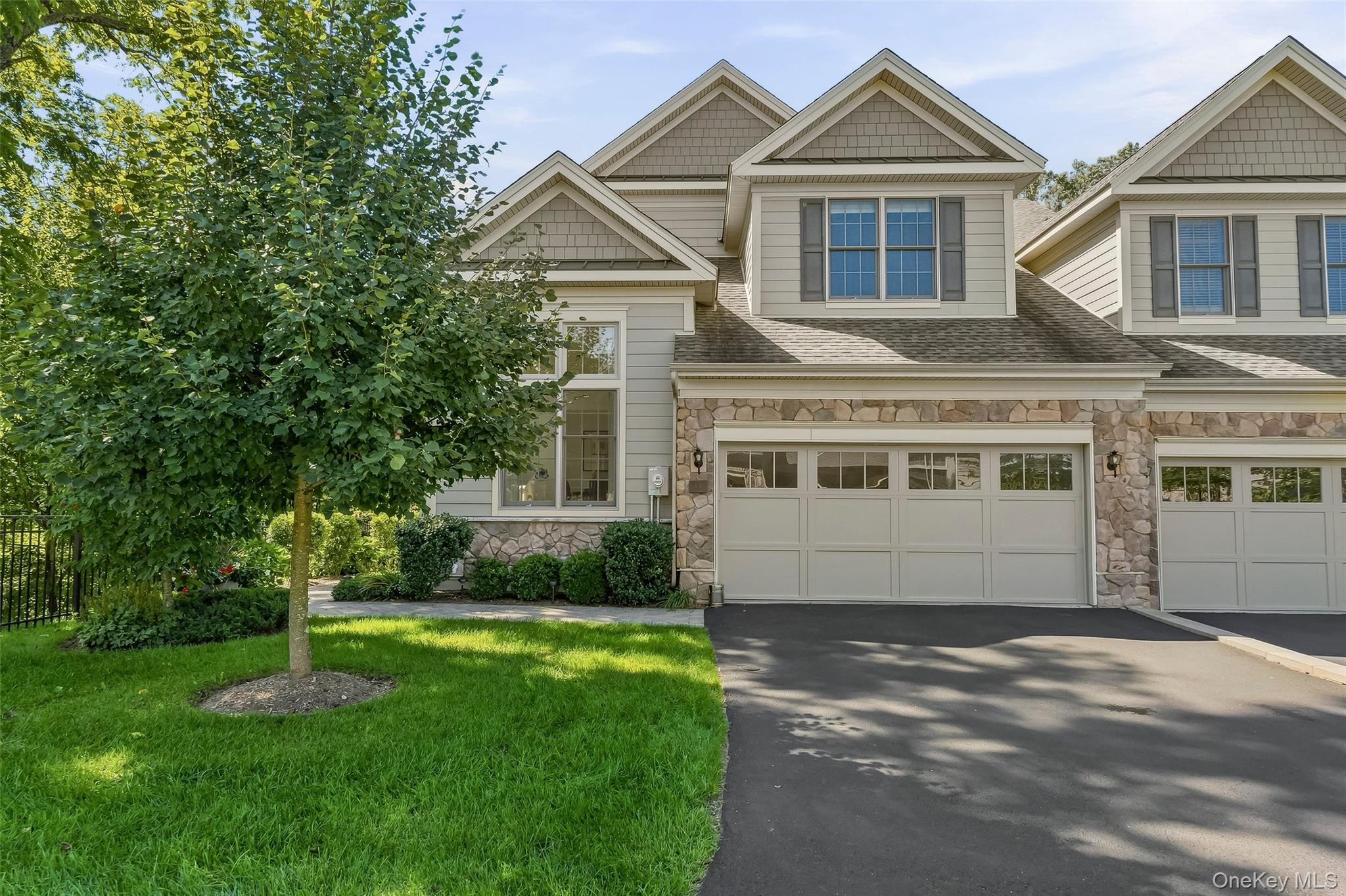 Large beige house with stone facade, two garage doors, and a leafy tree beside the driveway.