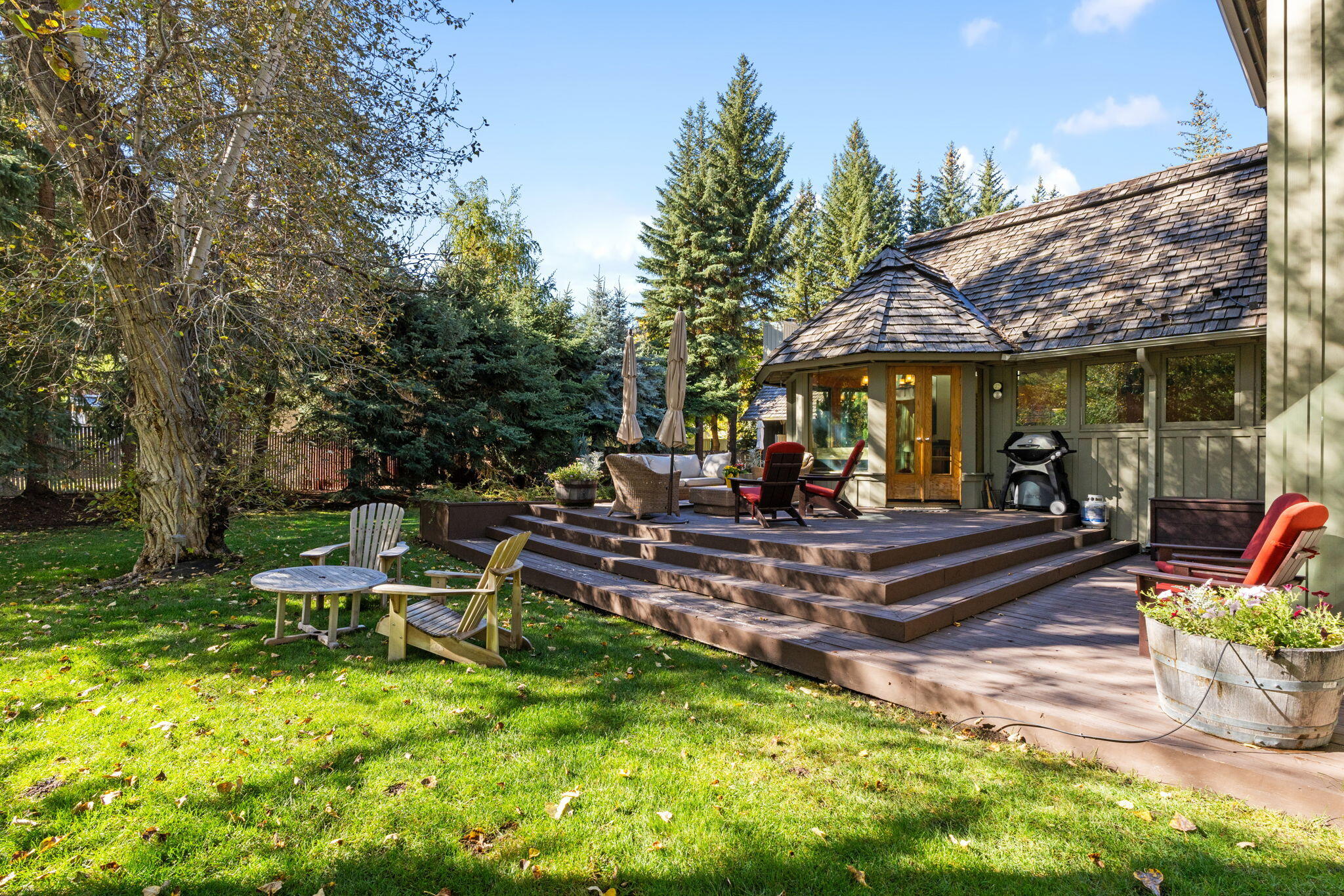 Home with a tiered furnished deck, grill, and leafy lawn beneath mature trees.