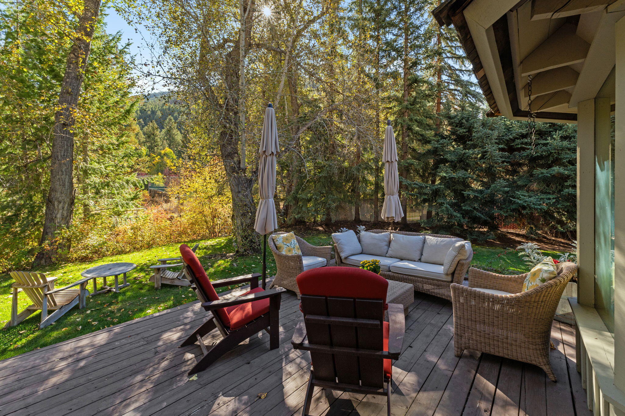 Furnished wooden deck with wicker seating and red Adirondack chairs overlooking an autumn woodland yard.