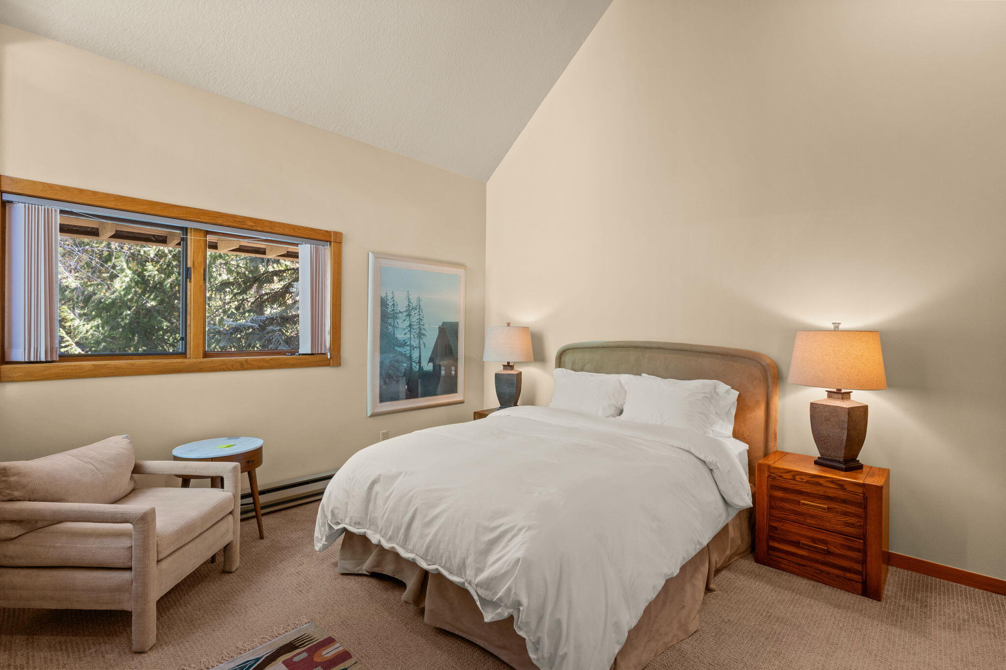 Bedroom with a king-size bed, vaulted ceiling, and wood-trimmed window overlooking evergreen trees.