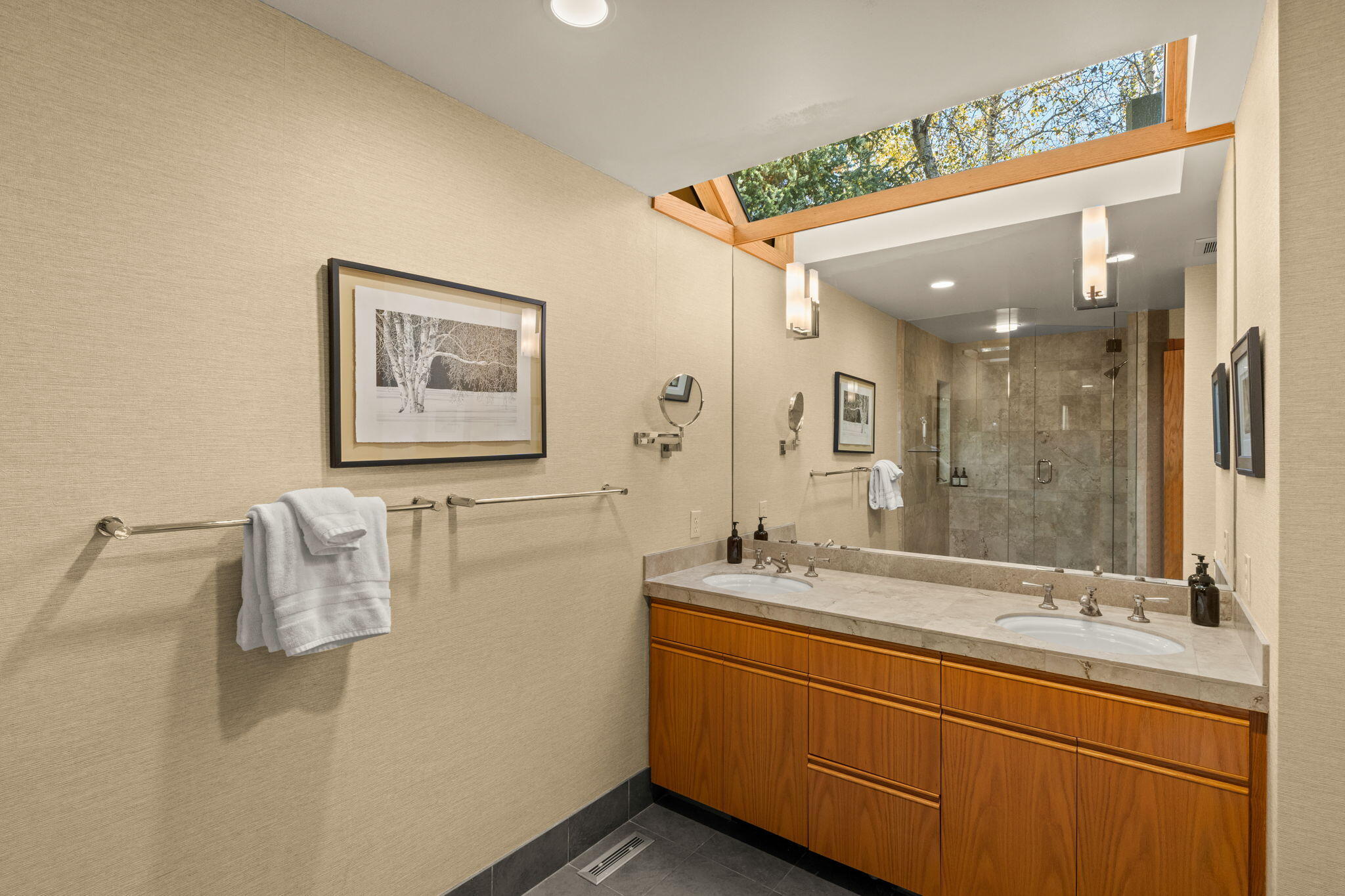 Bathroom with a double-sink wood vanity, glass shower, and wood-framed skylight.