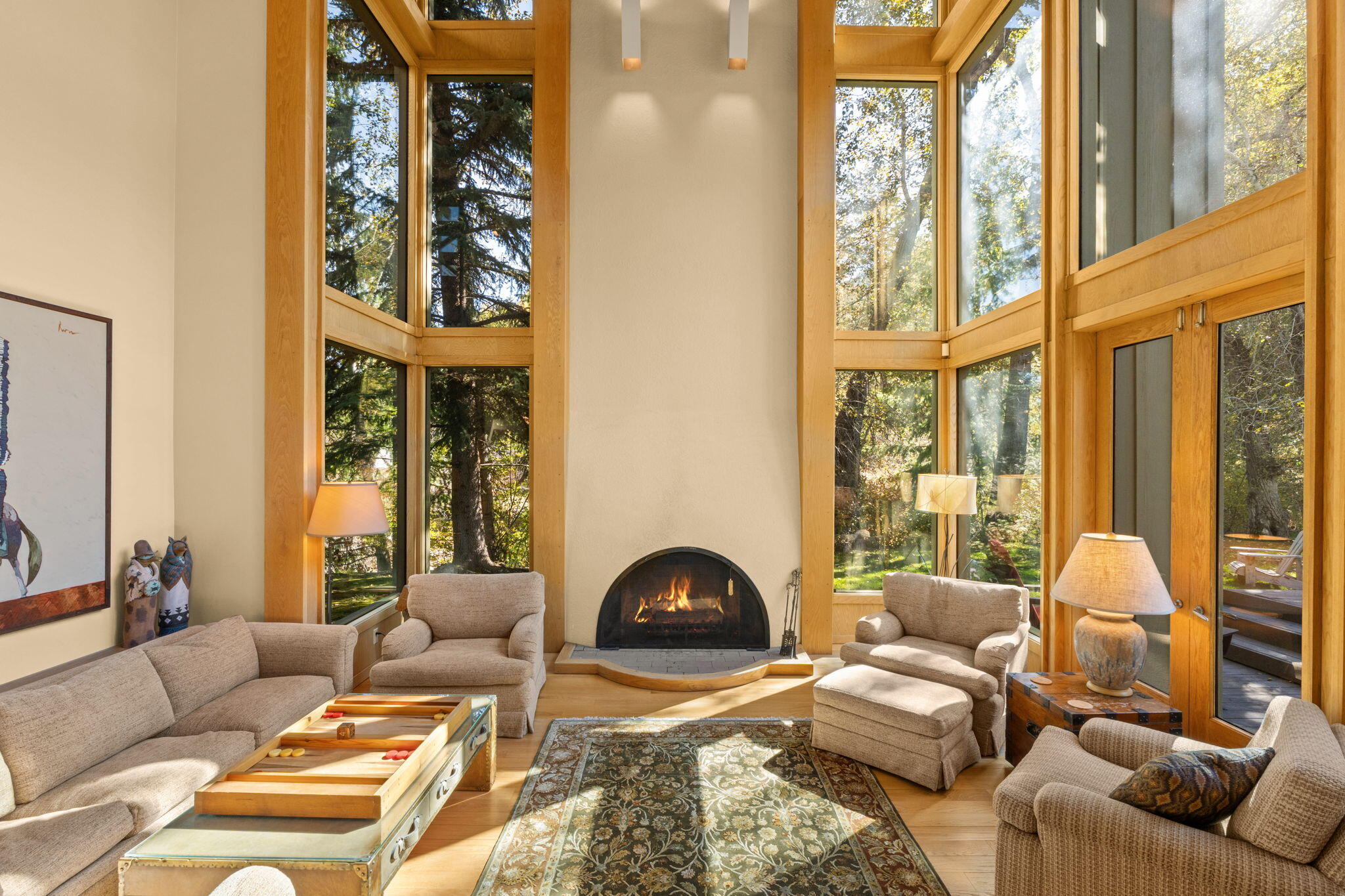 Living room with a tall arched fireplace, vaulted windows, and views of leafy trees.