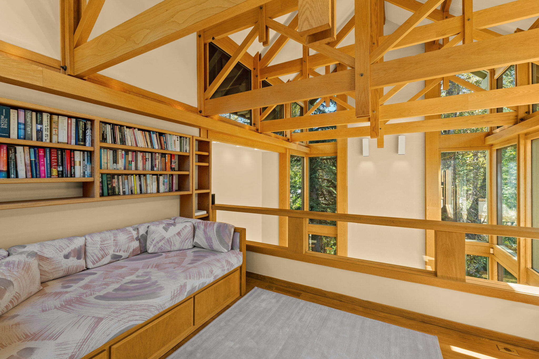 Loft sitting area with built-in cushioned bench, bookshelves, exposed timber beams, and tall windows overlooking trees.