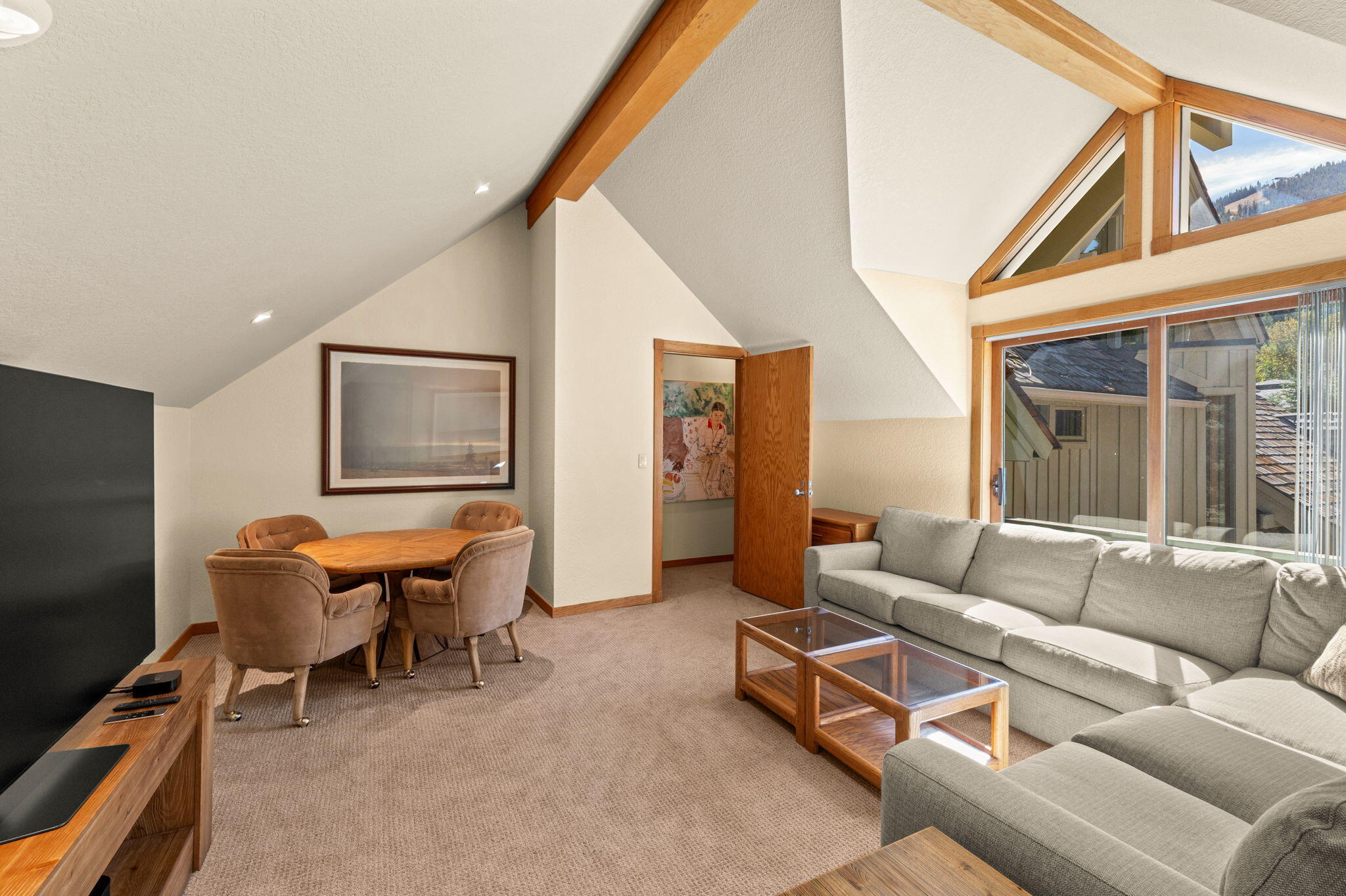 Living and dining room with vaulted wood-beamed ceiling, gray sectional, and large windows.