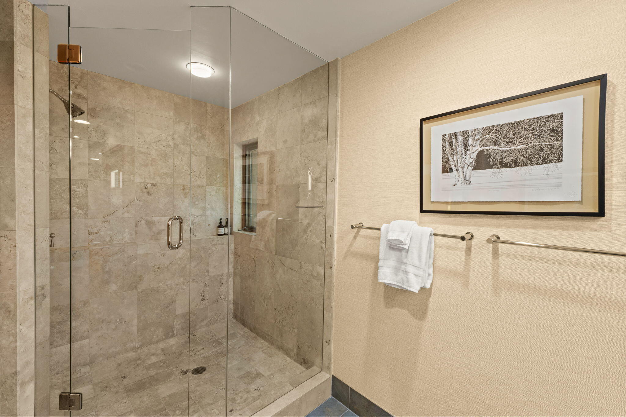 Stone-tiled walk-in shower with glass doors, white towels, and framed tree artwork.