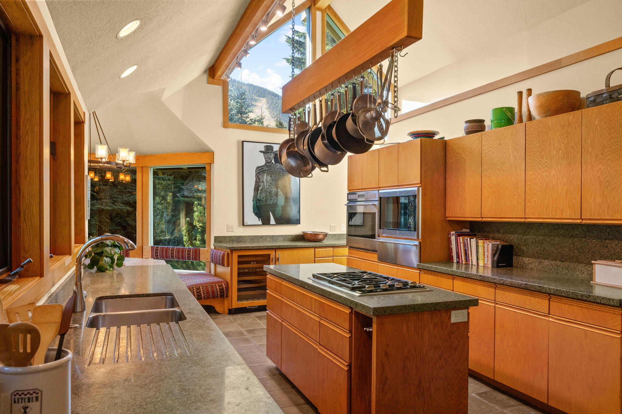 Kitchen with warm wood cabinetry, granite counters, hanging pots, and mountain views through high windows.