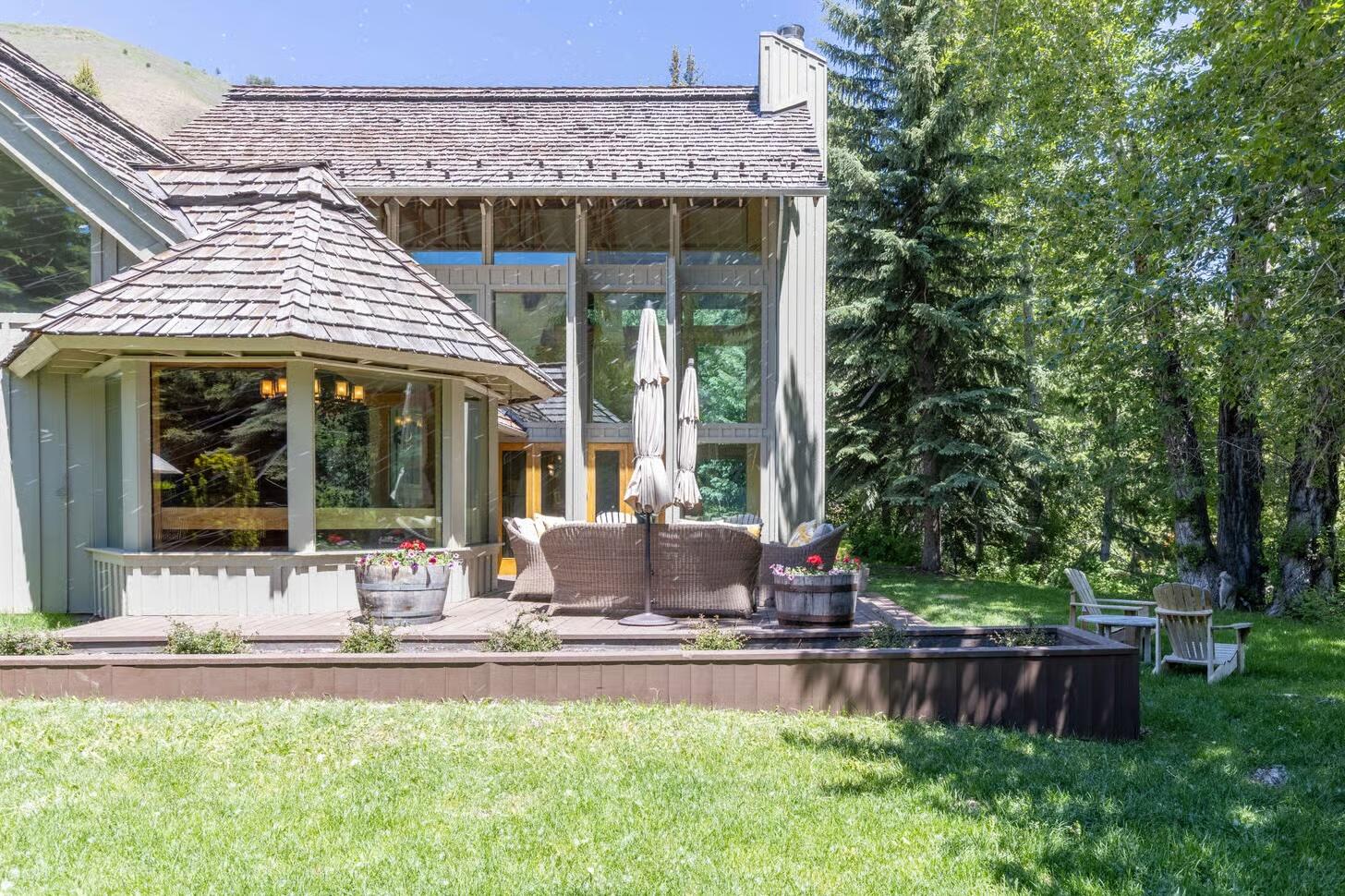 Home exterior with a raised deck, patio seating, and large windows surrounded by trees.
