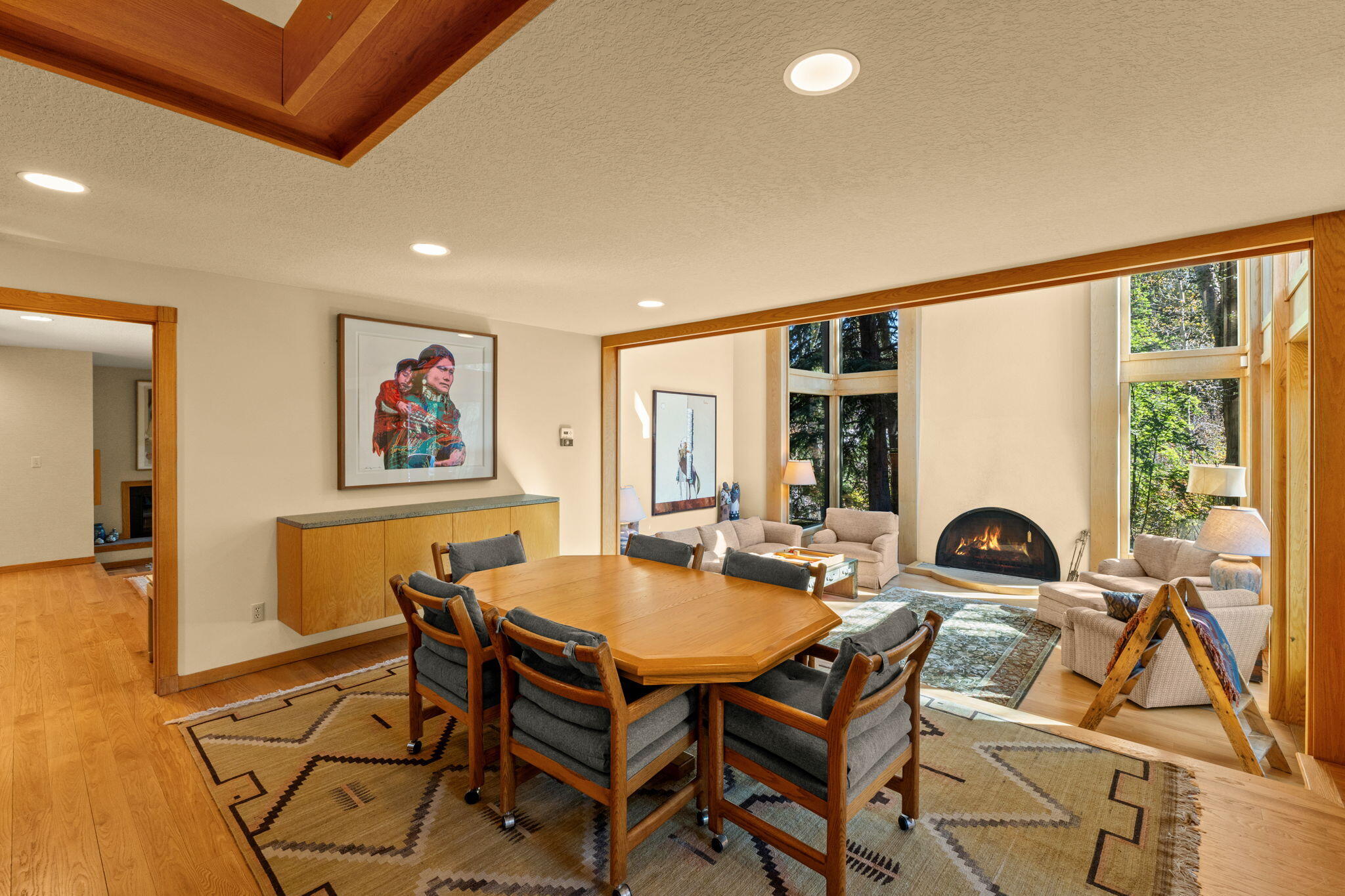 Open living and dining room with a large wooden table, tall windows, and a fireplace.