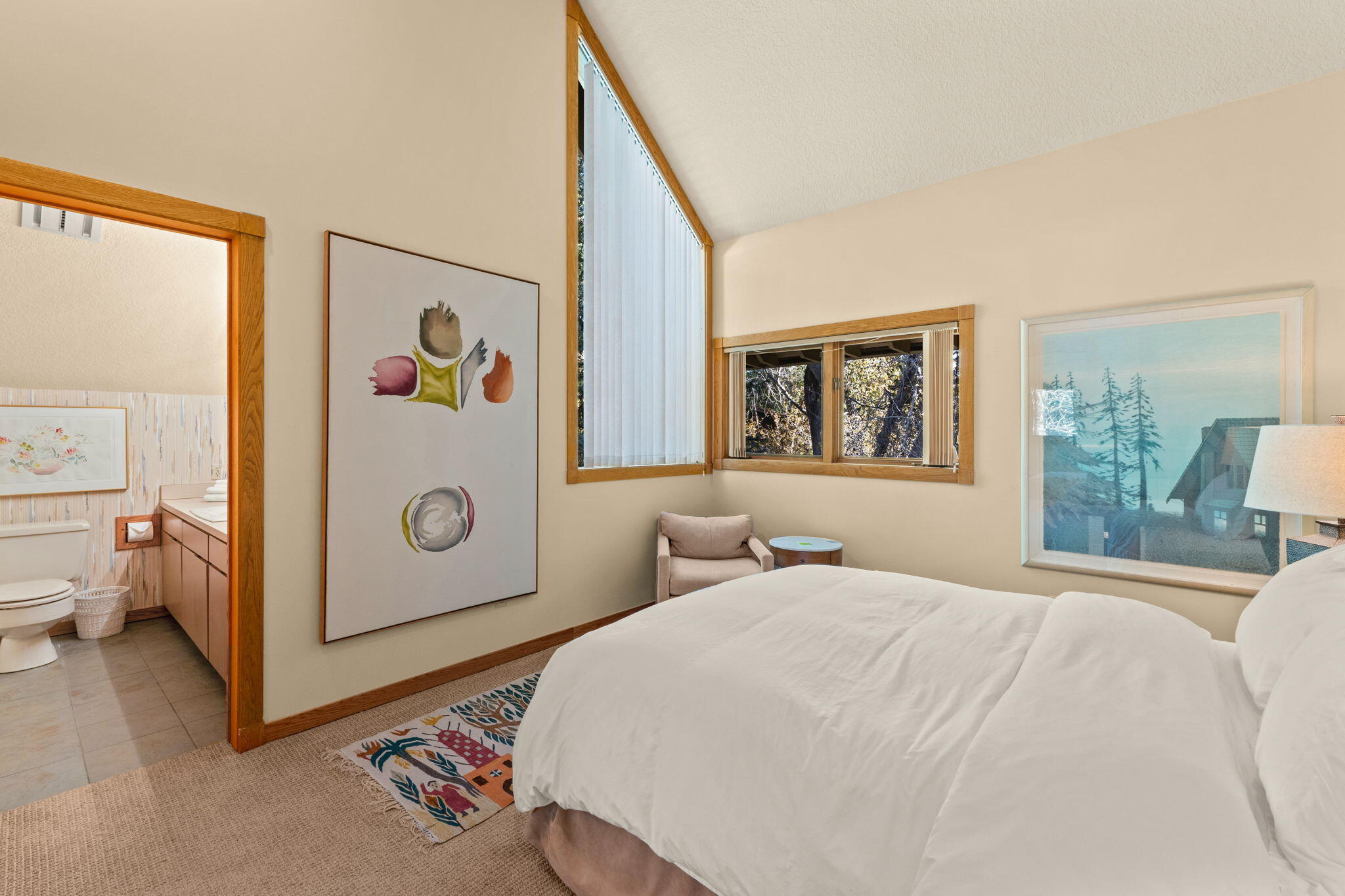 Bedroom with a white bed, colorful rug, wood-trimmed windows, and an adjoining bathroom.