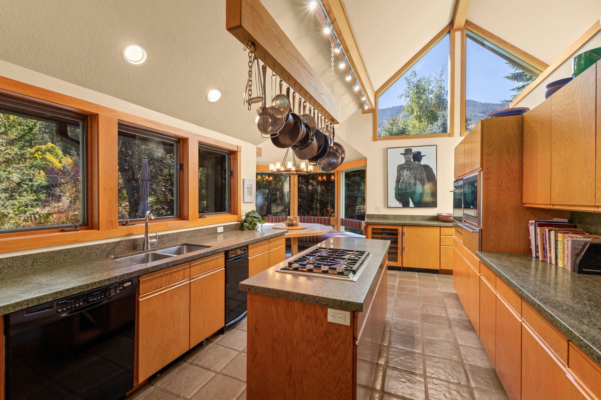Kitchen with wood cabinetry, a central cooktop island, hanging pots, and tall windows overlooking trees.