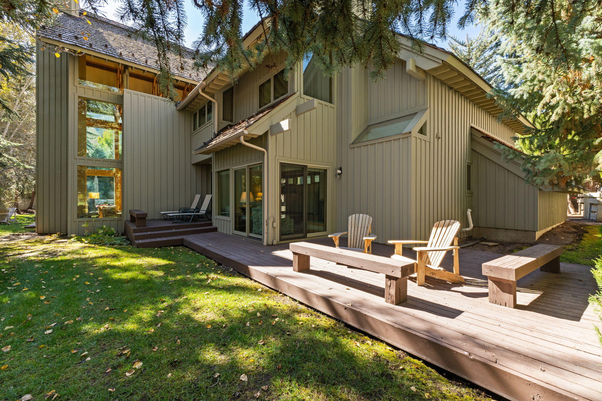 Home’s rear exterior with a broad wooden deck, Adirondack chairs, and evergreen trees.