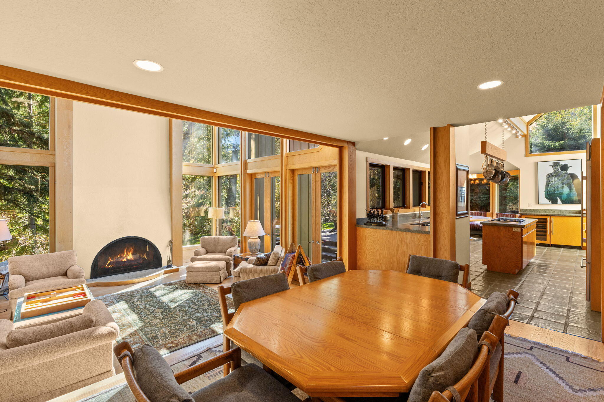 Open-plan living and dining area with a curved fireplace, large wood table, and forest-facing windows.