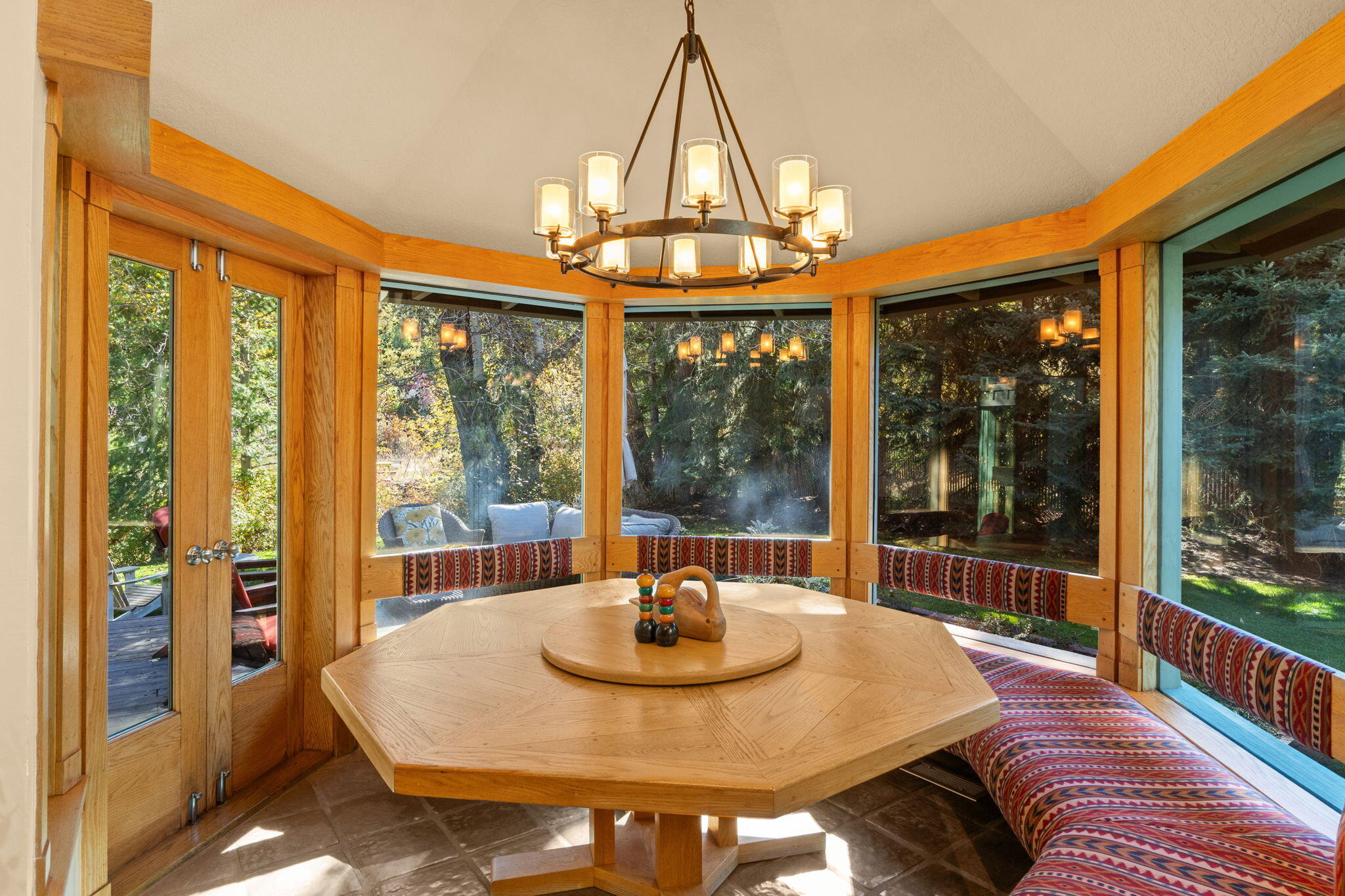 Dining nook with an octagonal wood table, patterned wraparound bench, and windows overlooking trees.