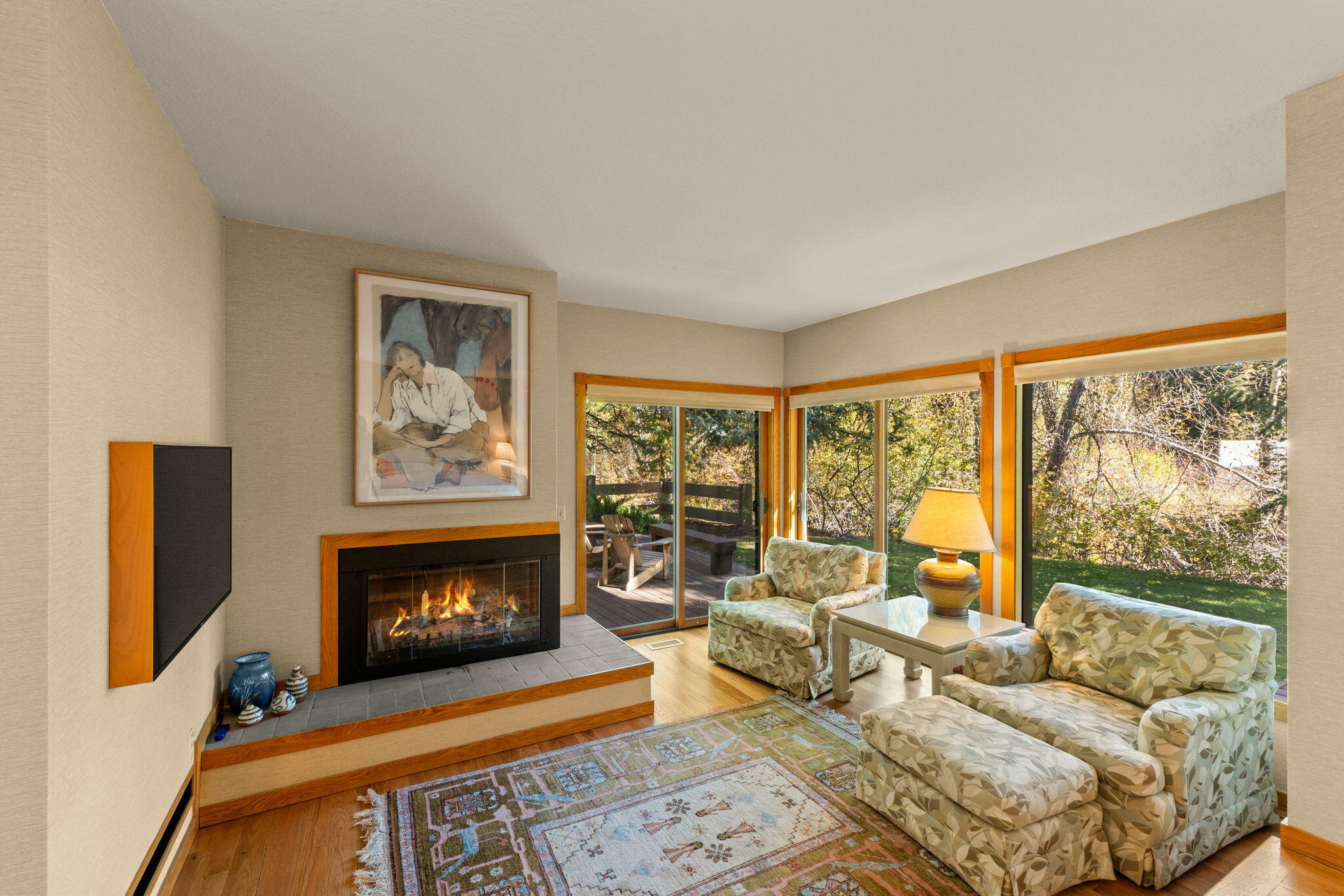 Living room with a lit fireplace, patterned armchairs, and large windows overlooking a leafy garden.