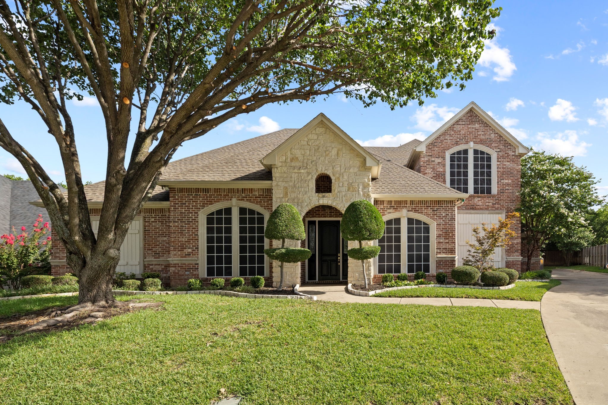 Two-story brick and stone home framed by a mature shade tree and landscaped lawn.