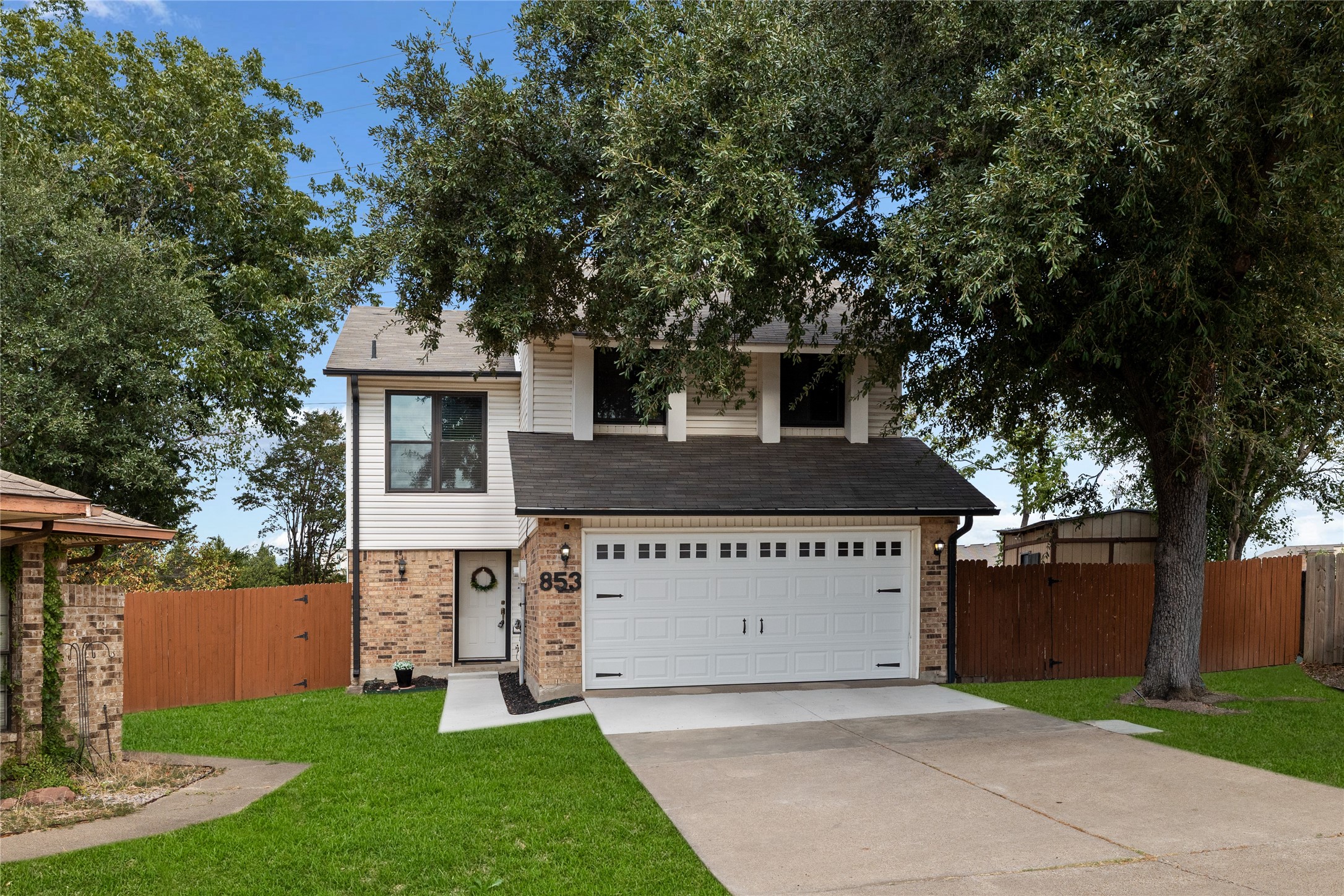 Two-story house with brick accents, a white double garage door, and a large shade tree in the front yard.