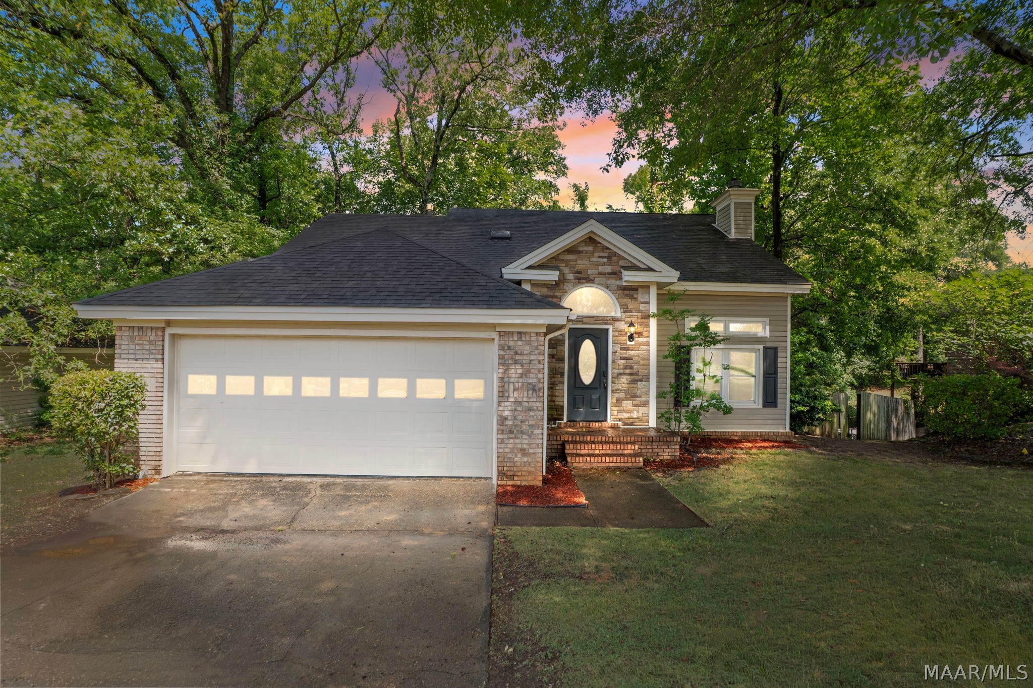 Single-story home with a two-car garage and stone-and-brick entry, framed by mature trees at dusk.