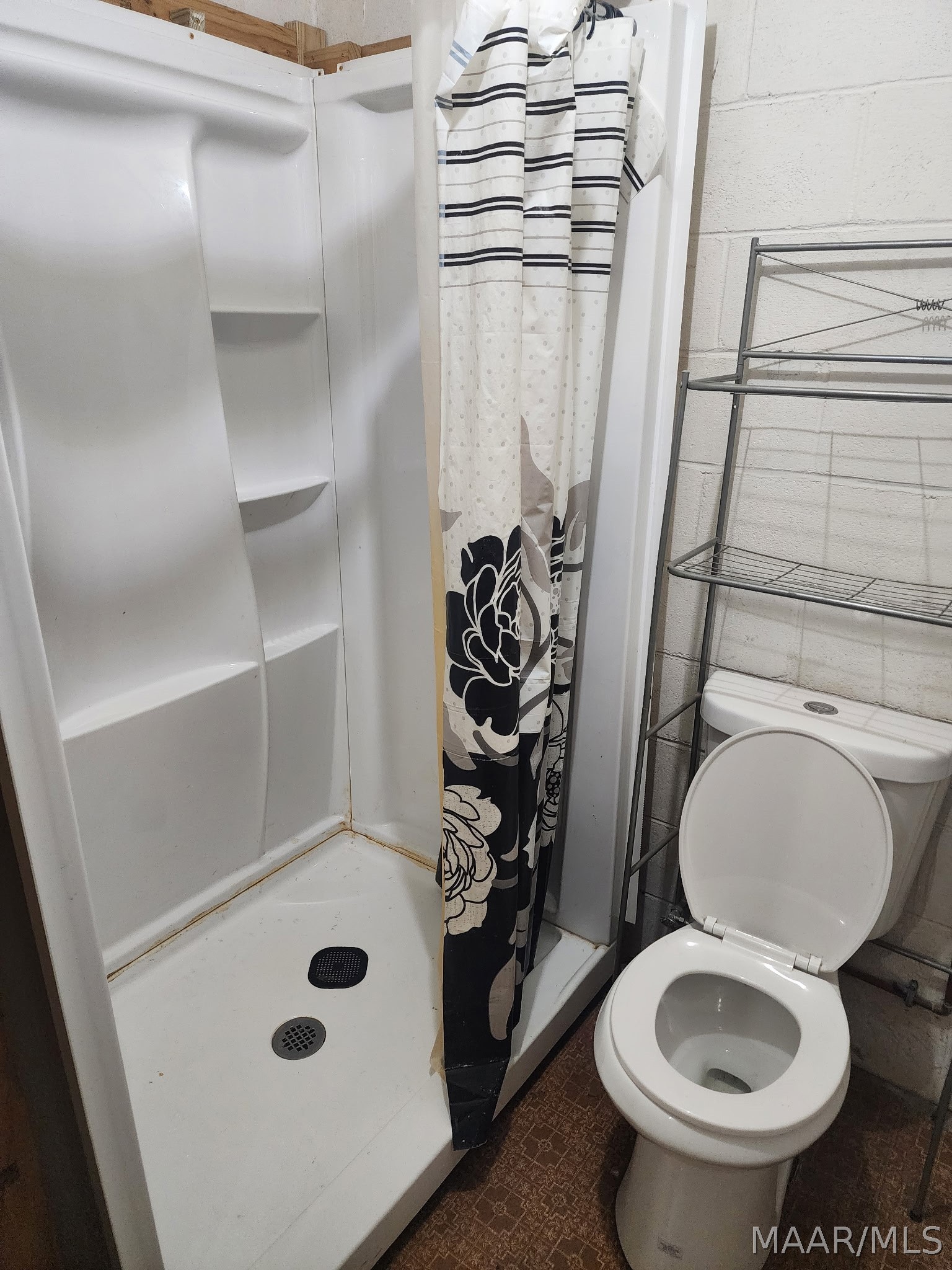 Compact bathroom with a white shower stall, patterned curtain, toilet, and metal storage rack.