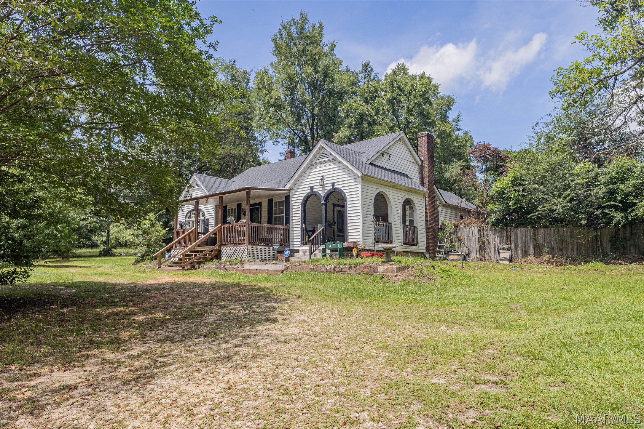 White house with a covered wooden porch, arched entry, brick chimney, and mature trees surrounding the yard.