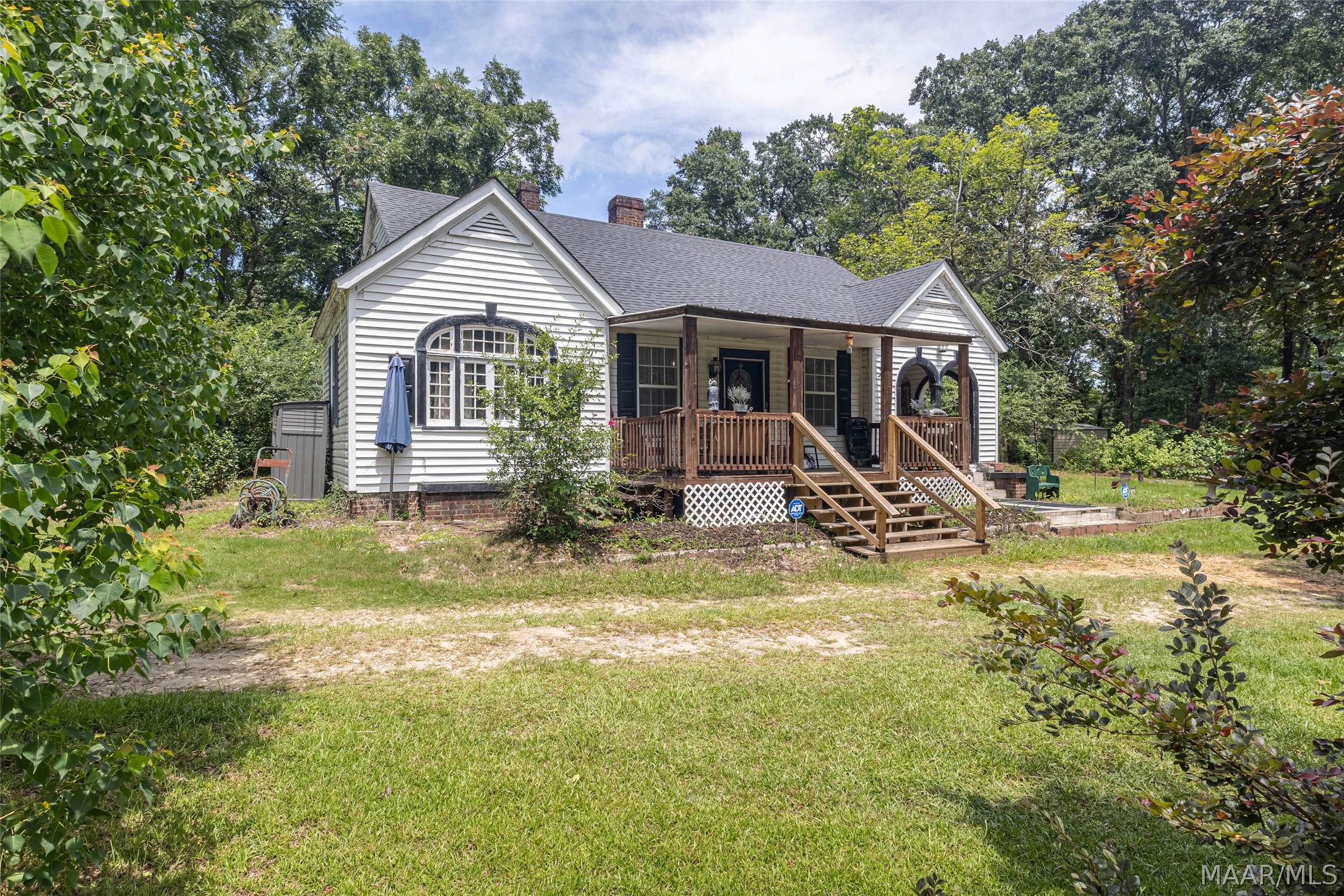 White-sided house with arched windows and a covered wooden porch, surrounded by trees and lawn.