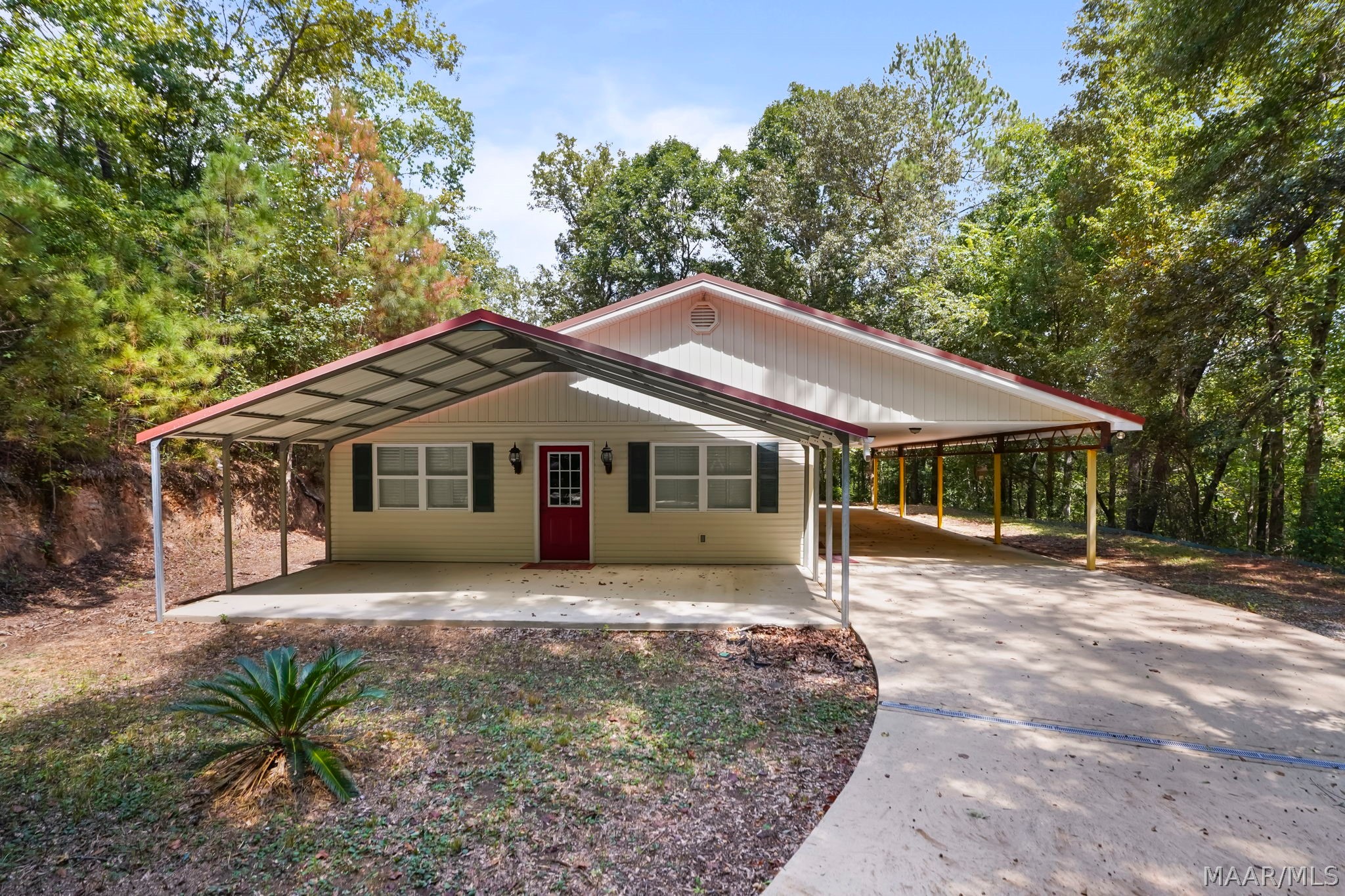Beige single-story home with a red door, large carport, and concrete driveway among trees.