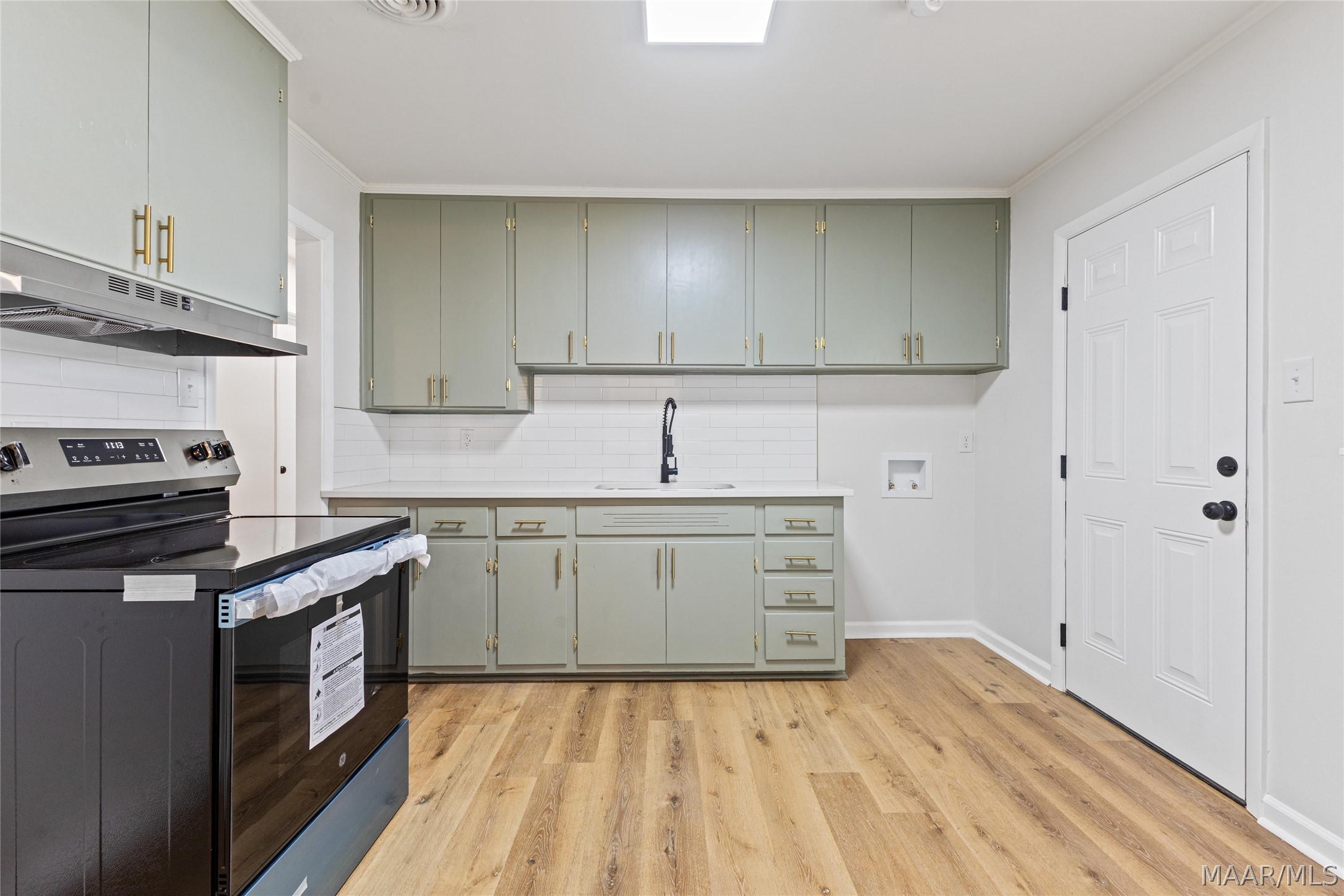 Vacant kitchen with sage-green cabinets, brass hardware, white tile backsplash, and light wood-look flooring.