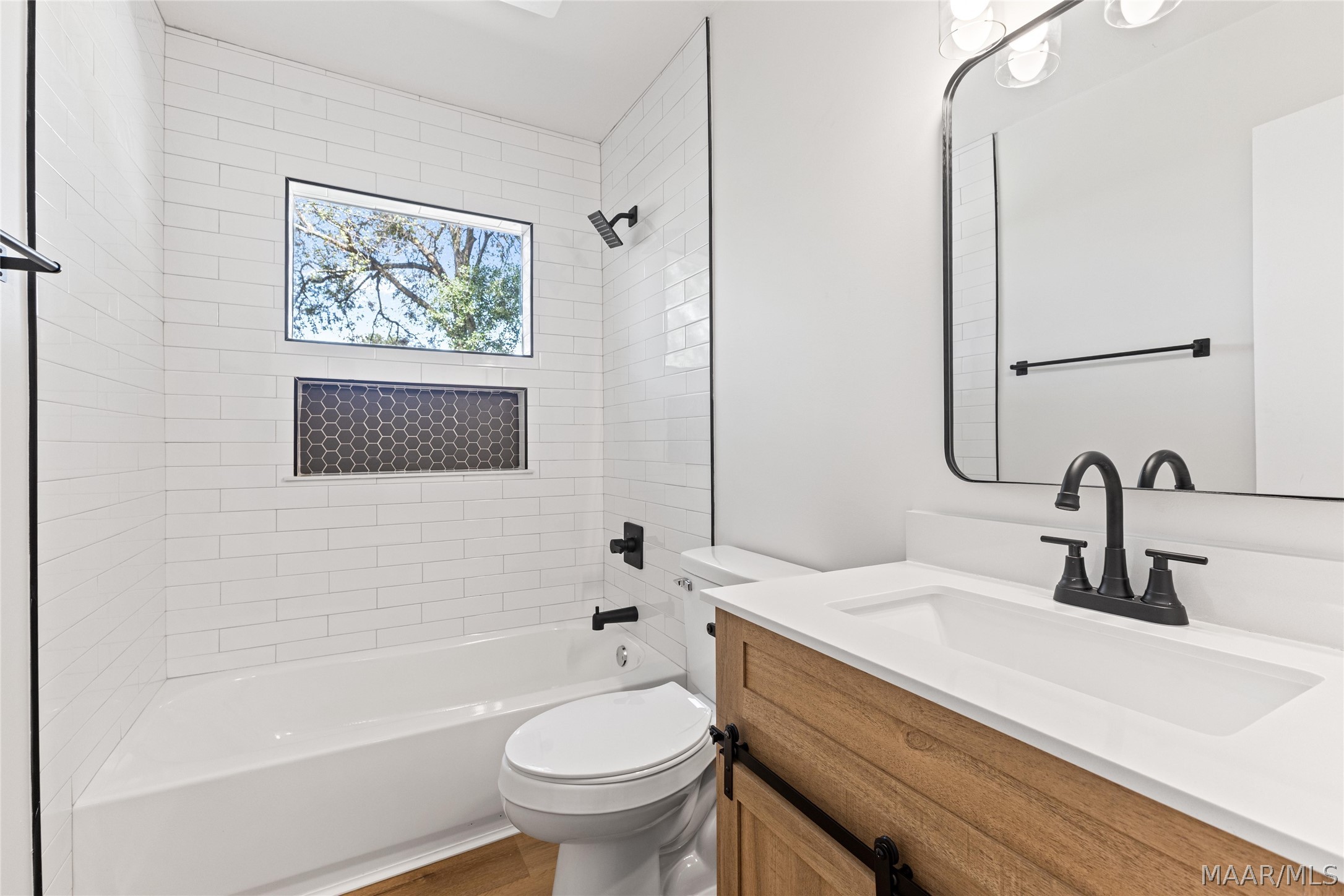 Bathroom with a tiled tub and shower, high window, and wood vanity with black fixtures.
