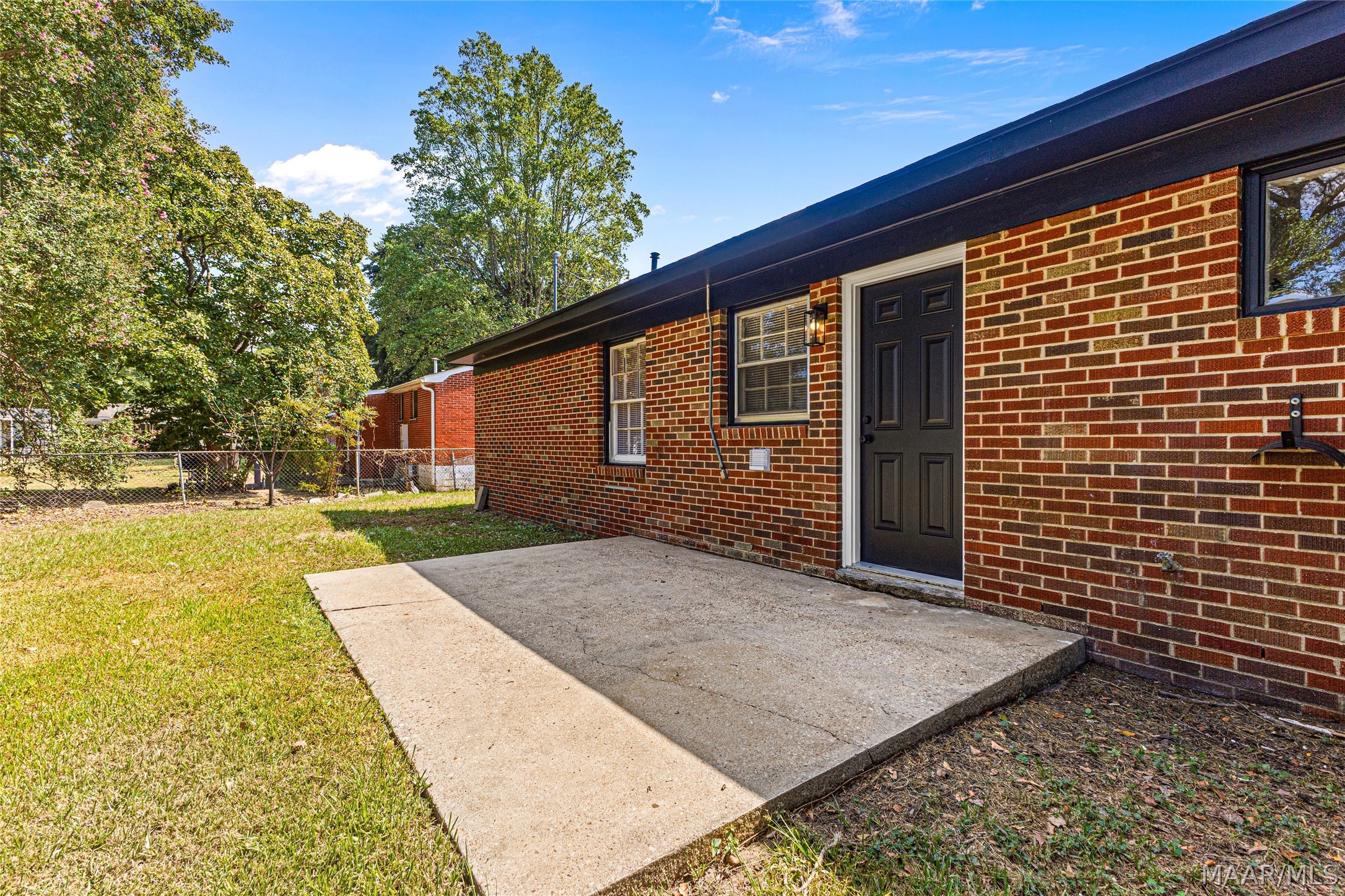 Red brick home’s black side entrance opens onto a broad concrete patio beside the lawn.