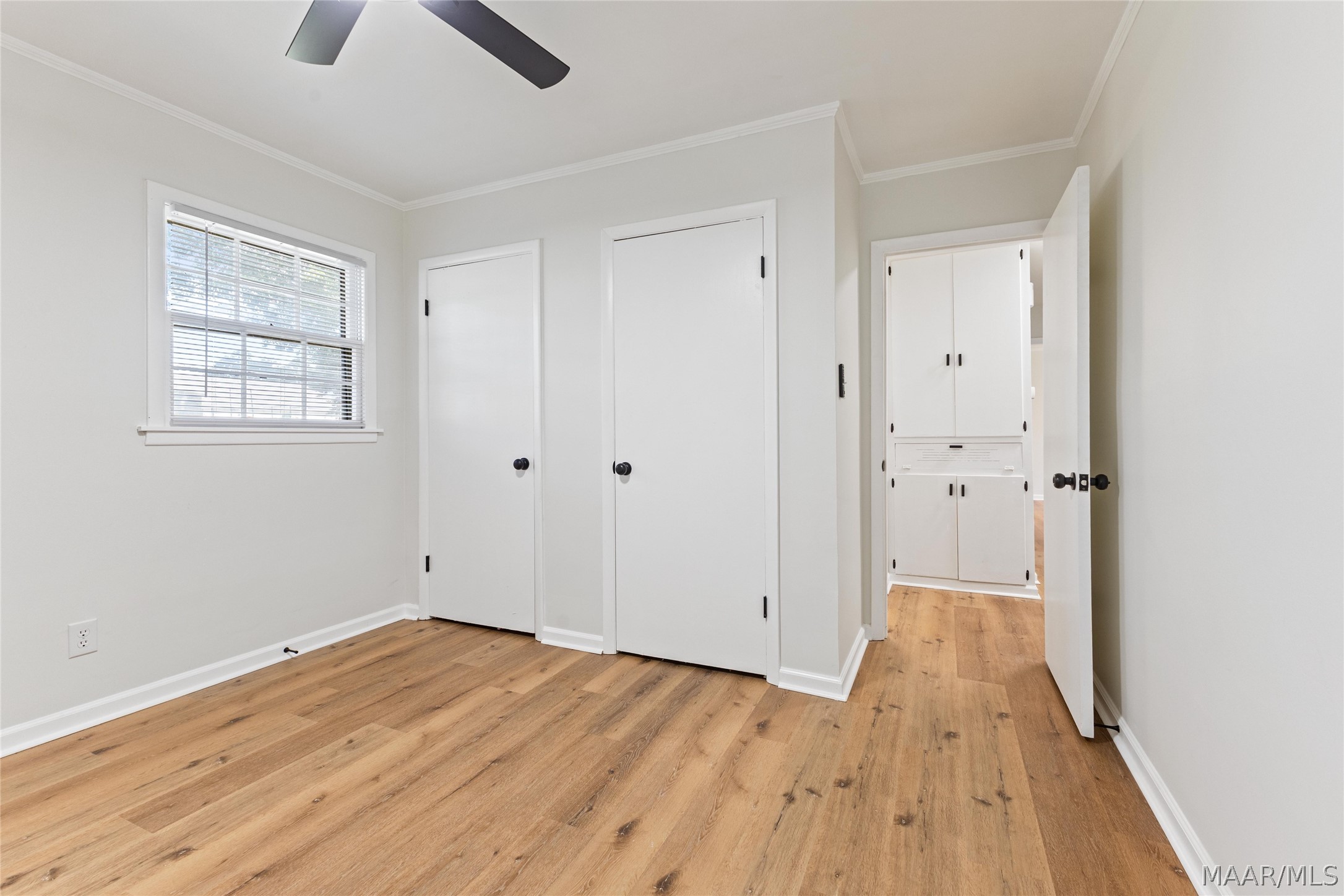 Empty room with warm wood flooring, a window with blinds, and built-in cabinets through the doorway.