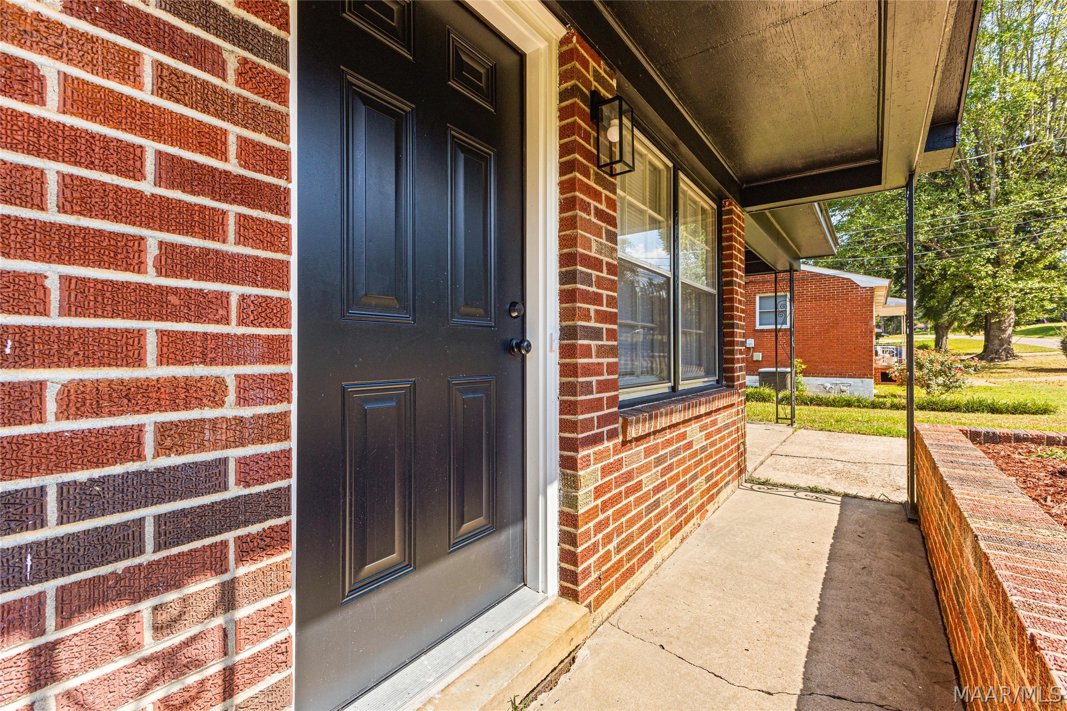 Brick home’s covered front porch with a black entry door, large windows, and concrete walkway.