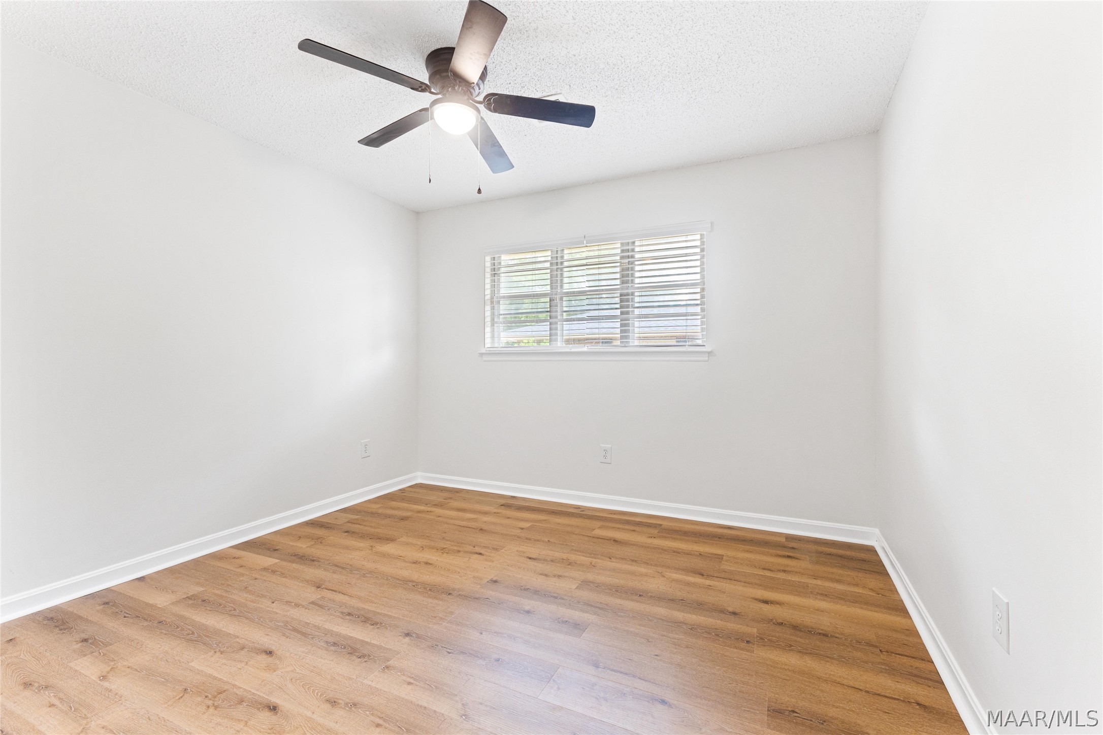 Empty room with warm wood floors, a ceiling fan, and a window with blinds.