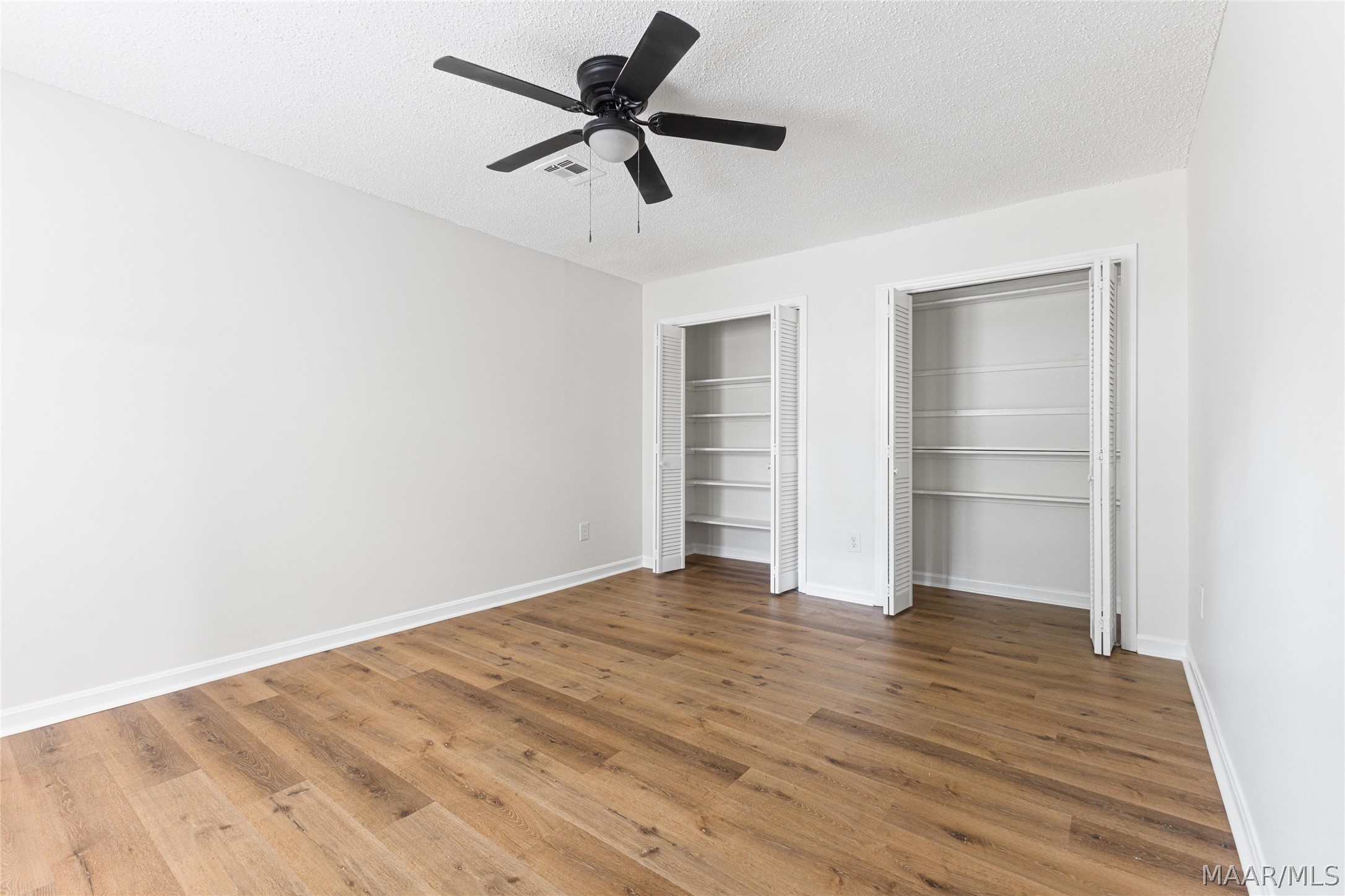 Empty bedroom with wood flooring, a black ceiling fan, and two open closets with built-in shelves.