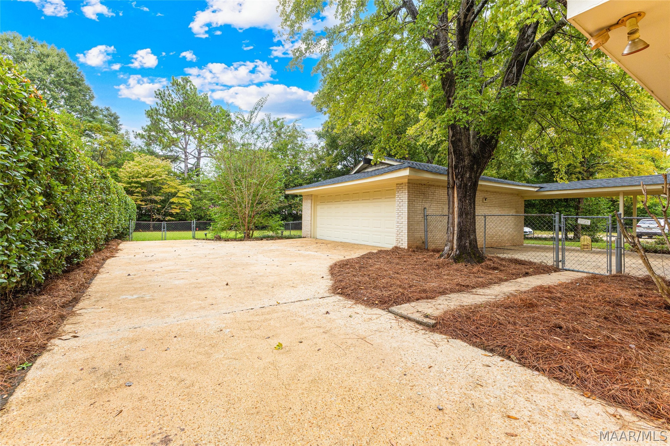 Brick garage and covered carport at the end of a wide driveway, shaded by mature trees.