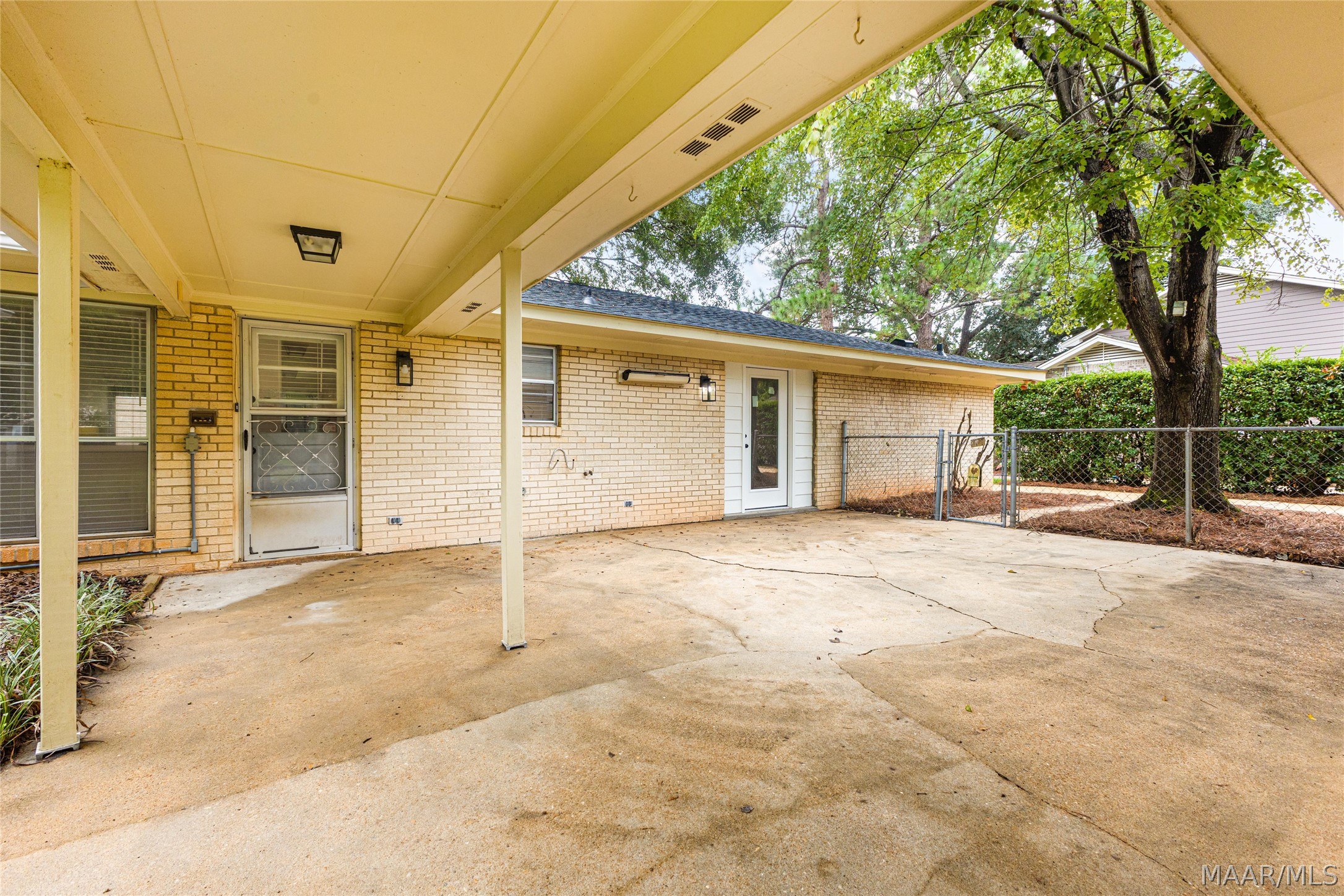 Covered concrete patio beside a pale brick house, with a fenced yard and mature trees.