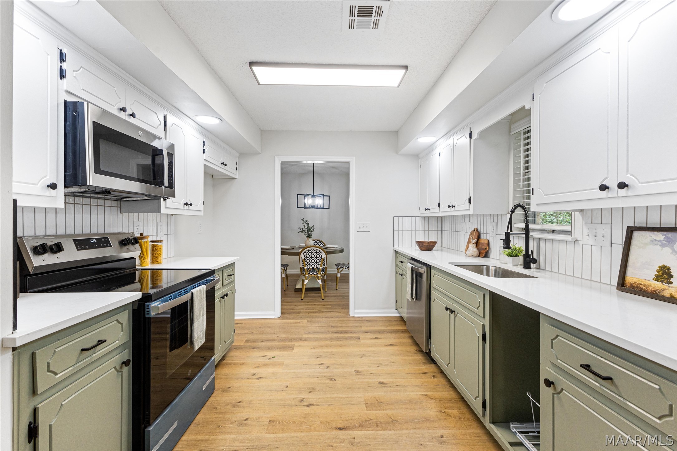 Galley kitchen with sage-green lower cabinets, stainless appliances, white counters, and warm wood floors.