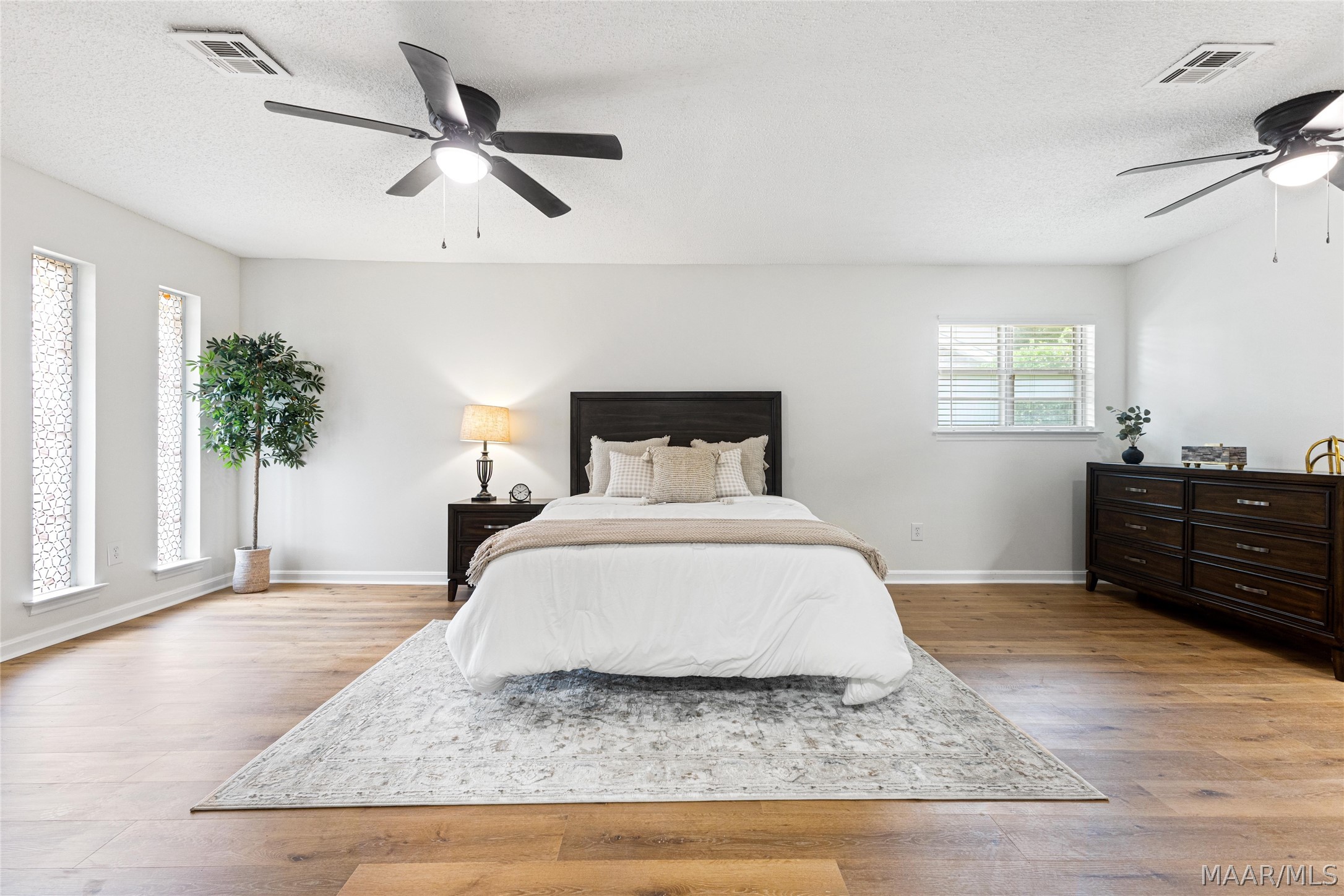 Staged bedroom with a centered bed, dark wood furniture, ceiling fans, and warm wood flooring.