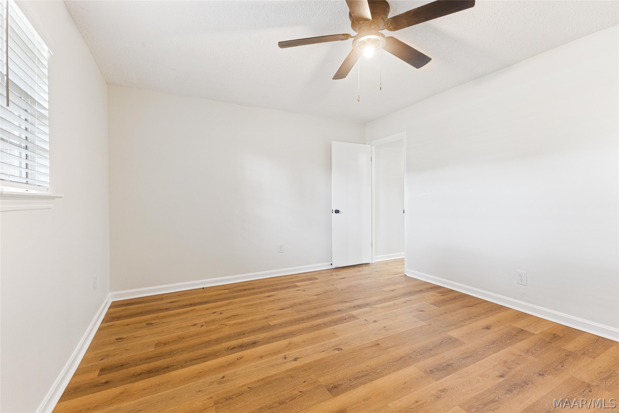 Empty bedroom with warm wood flooring, a ceiling fan, and a window with blinds.
