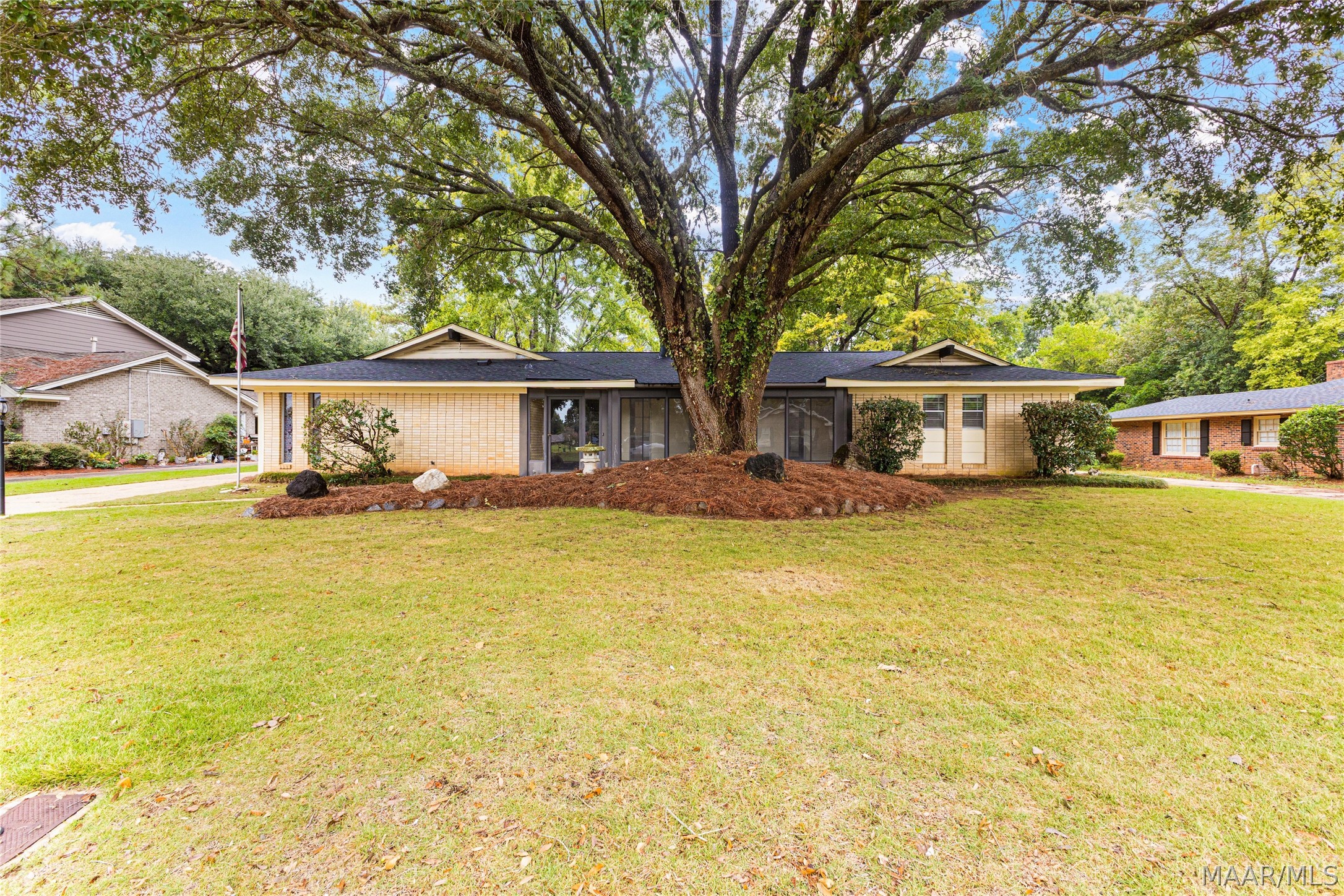 Single-story brick home beneath a large mature tree, with a broad front lawn.