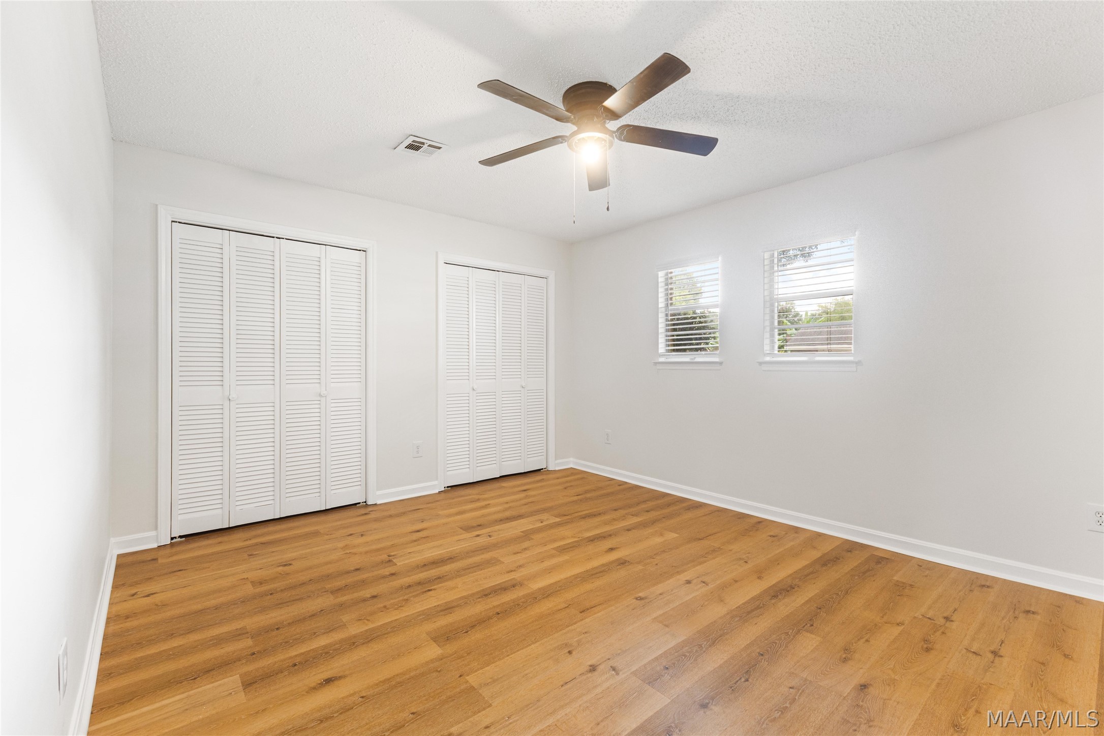 Empty bedroom with wood floors, louvered closets, two windows, and a ceiling fan.