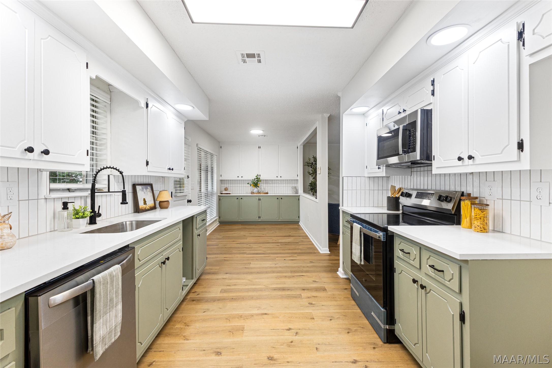 Kitchen with sage-green cabinets, white counters, stainless steel appliances, and warm wood floors.