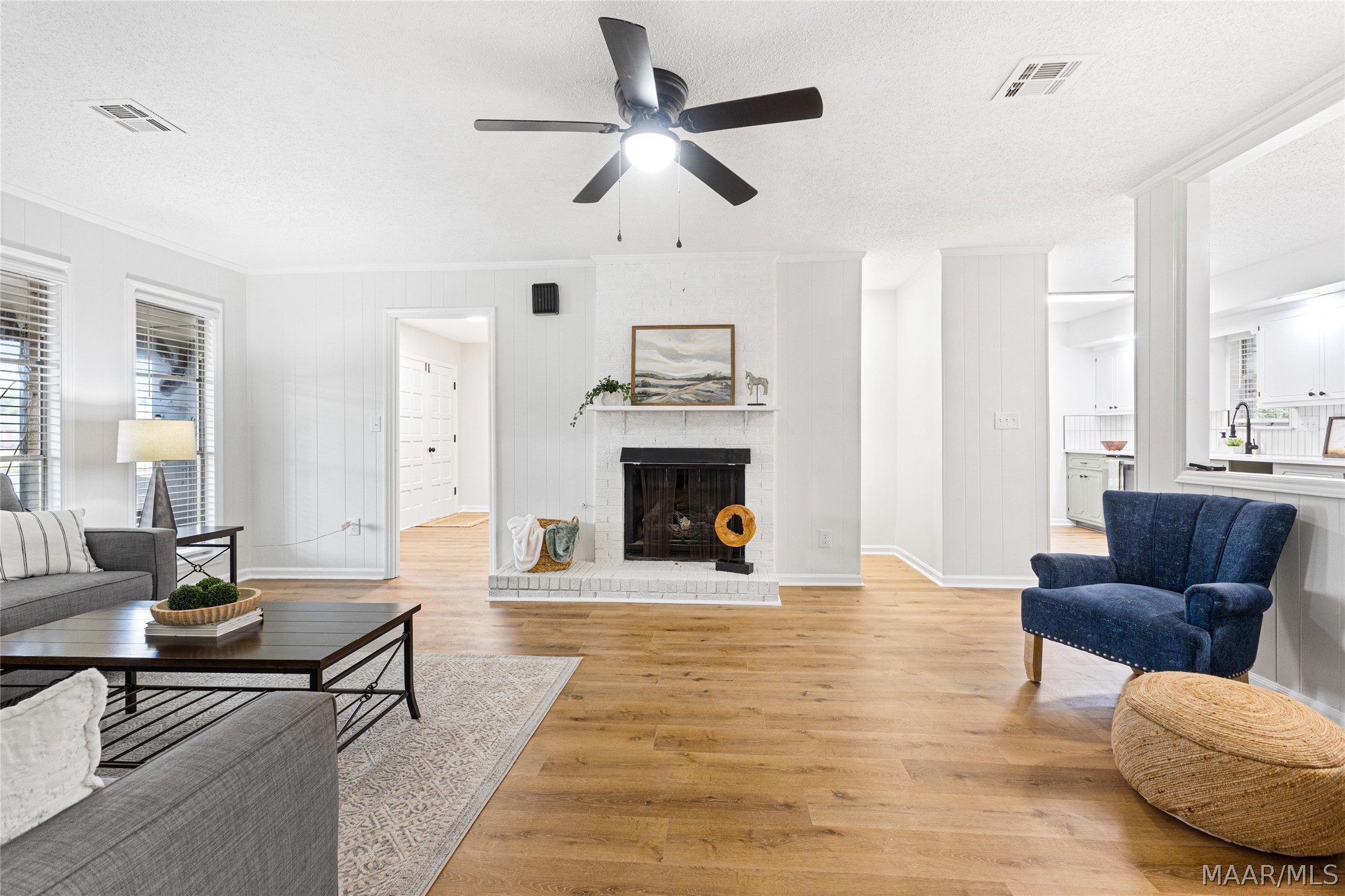 Living room with a white brick fireplace, wood floors, and an opening to the kitchen.