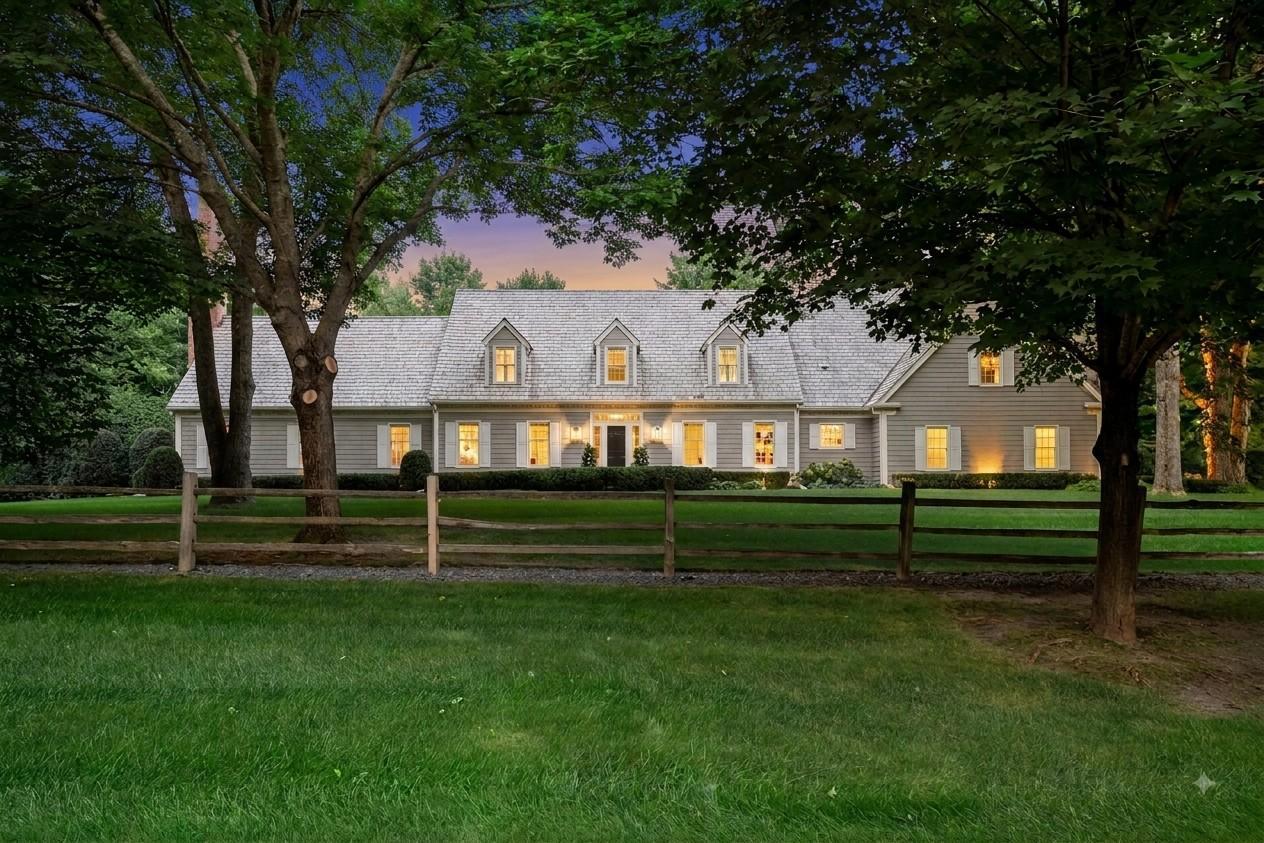 Gray-shingled house with glowing windows behind a wooden fence, framed by mature trees at dusk.