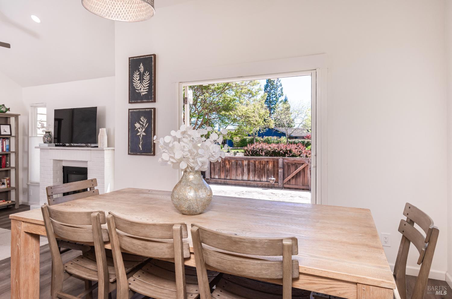 Dining area with a large wooden table, white brick fireplace, and open door to a leafy fenced yard.