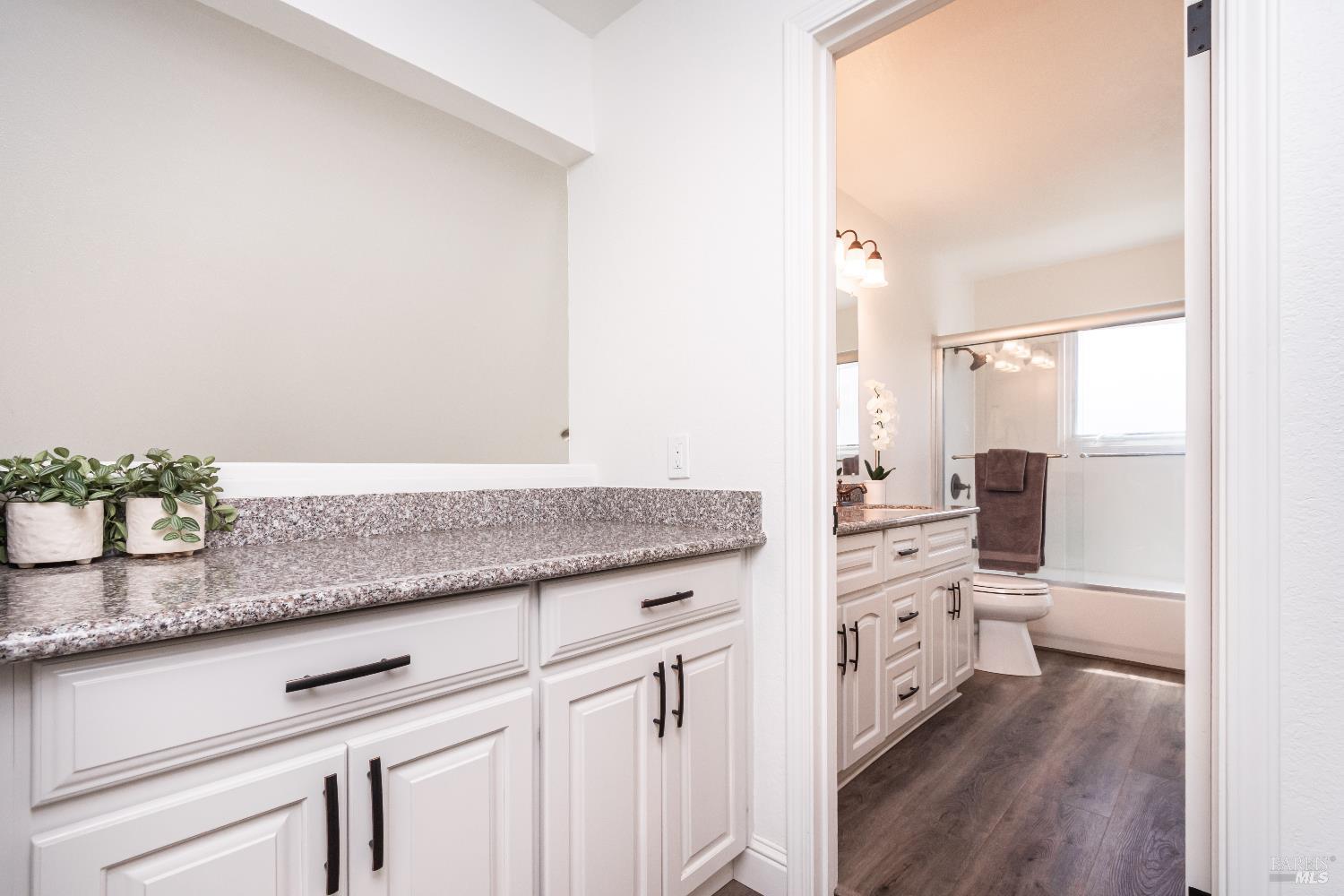 Bathroom vanity with granite counters and white cabinetry, looking through to a tub and toilet.