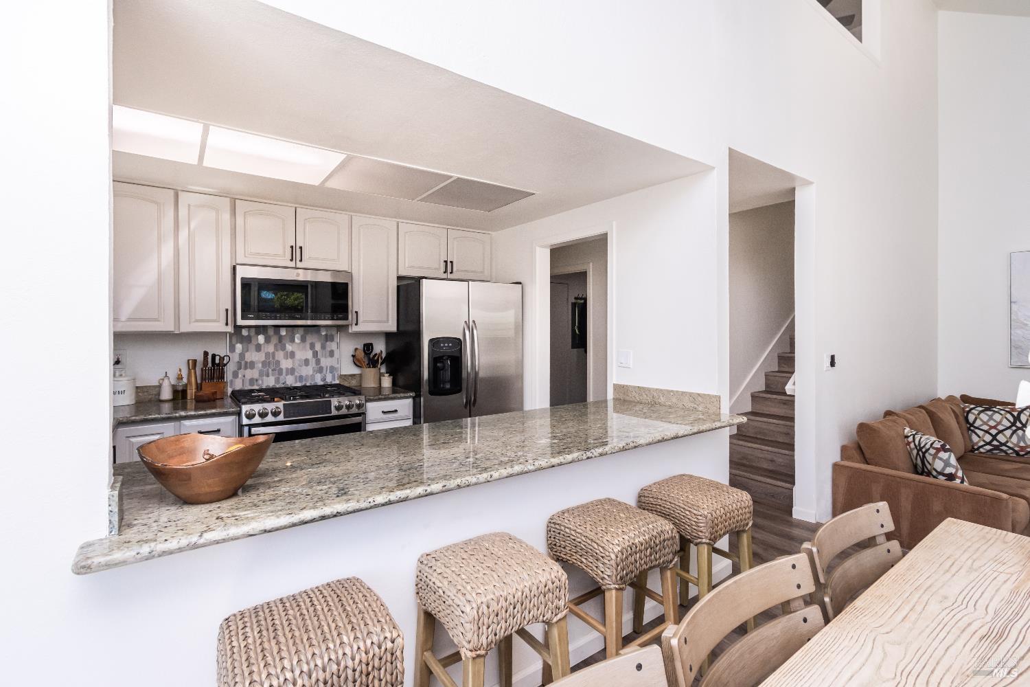 Kitchen with a long granite breakfast bar, woven stools, white cabinetry, and stainless steel appliances.