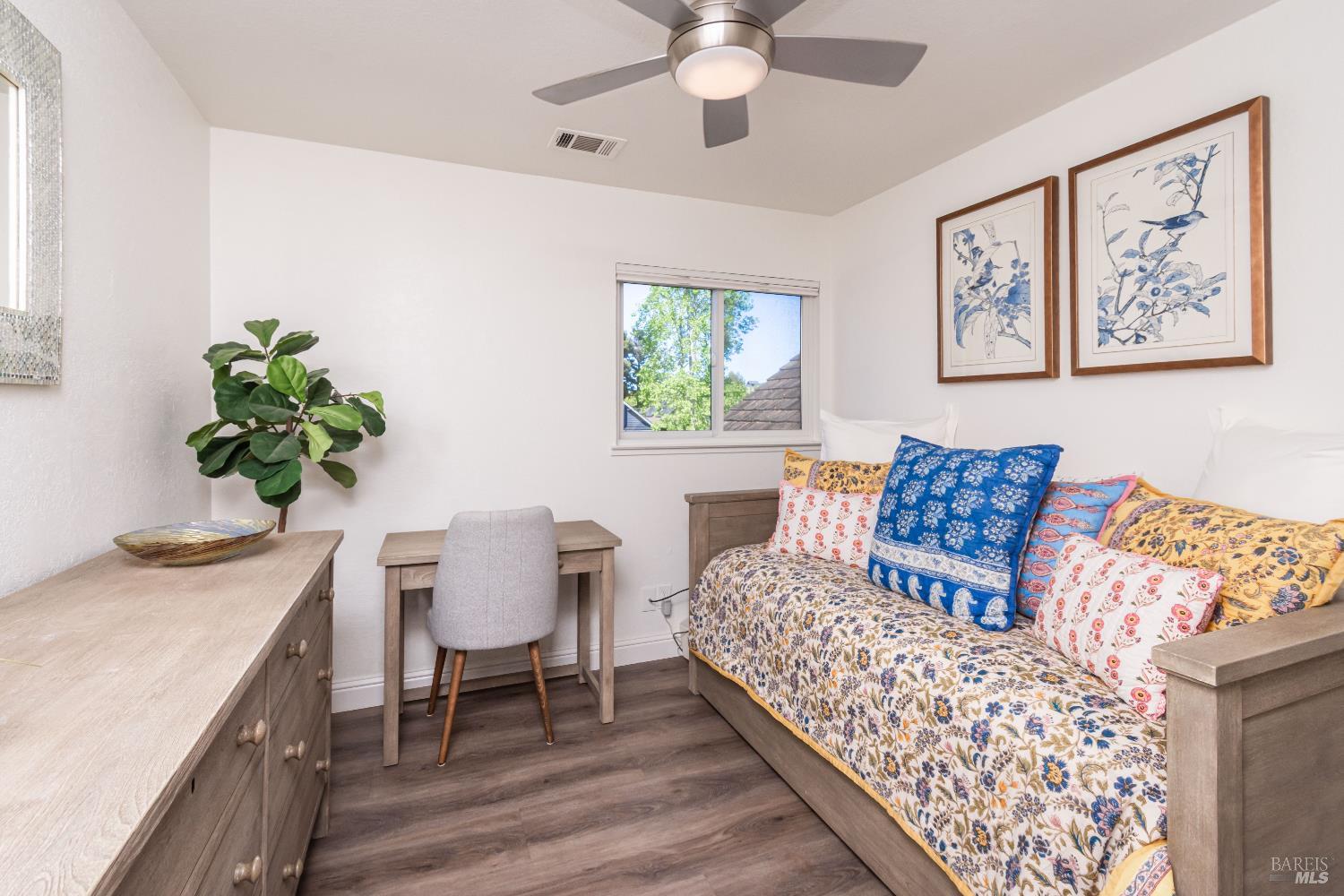 Bedroom with a floral-covered bed, colorful pillows, a small desk, and framed botanical prints.