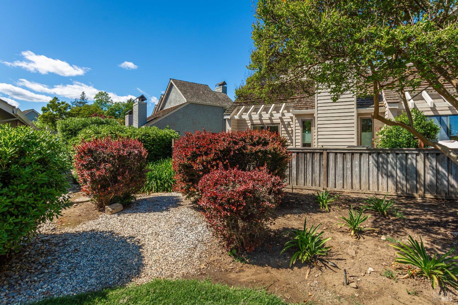 House exterior behind a wooden fence, with red shrubs and a gravel path.